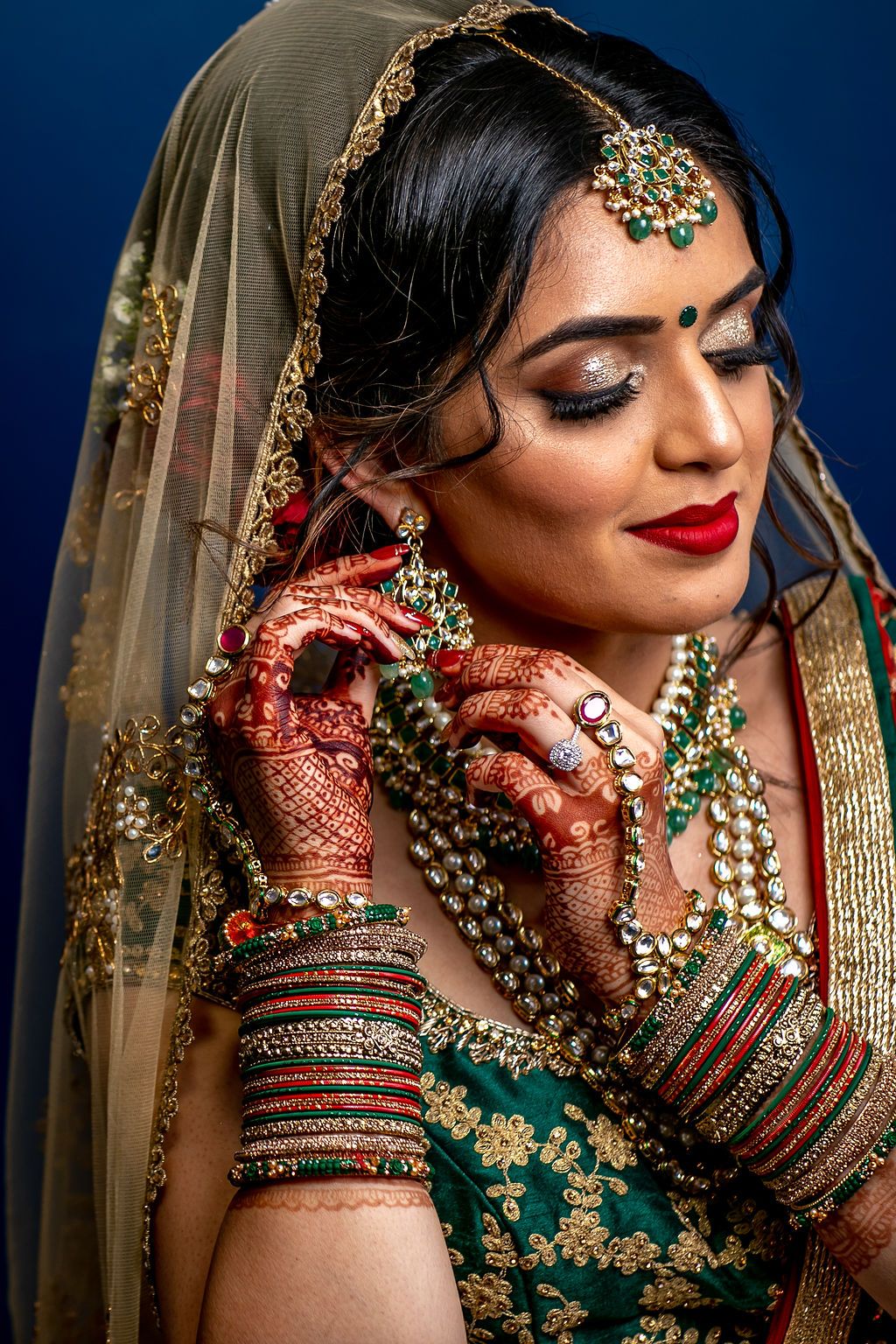 Indian bride in green lehenga with gold embroidery, mehndi hands, kundan jewelry, maang tikka, and red lips on blue background.