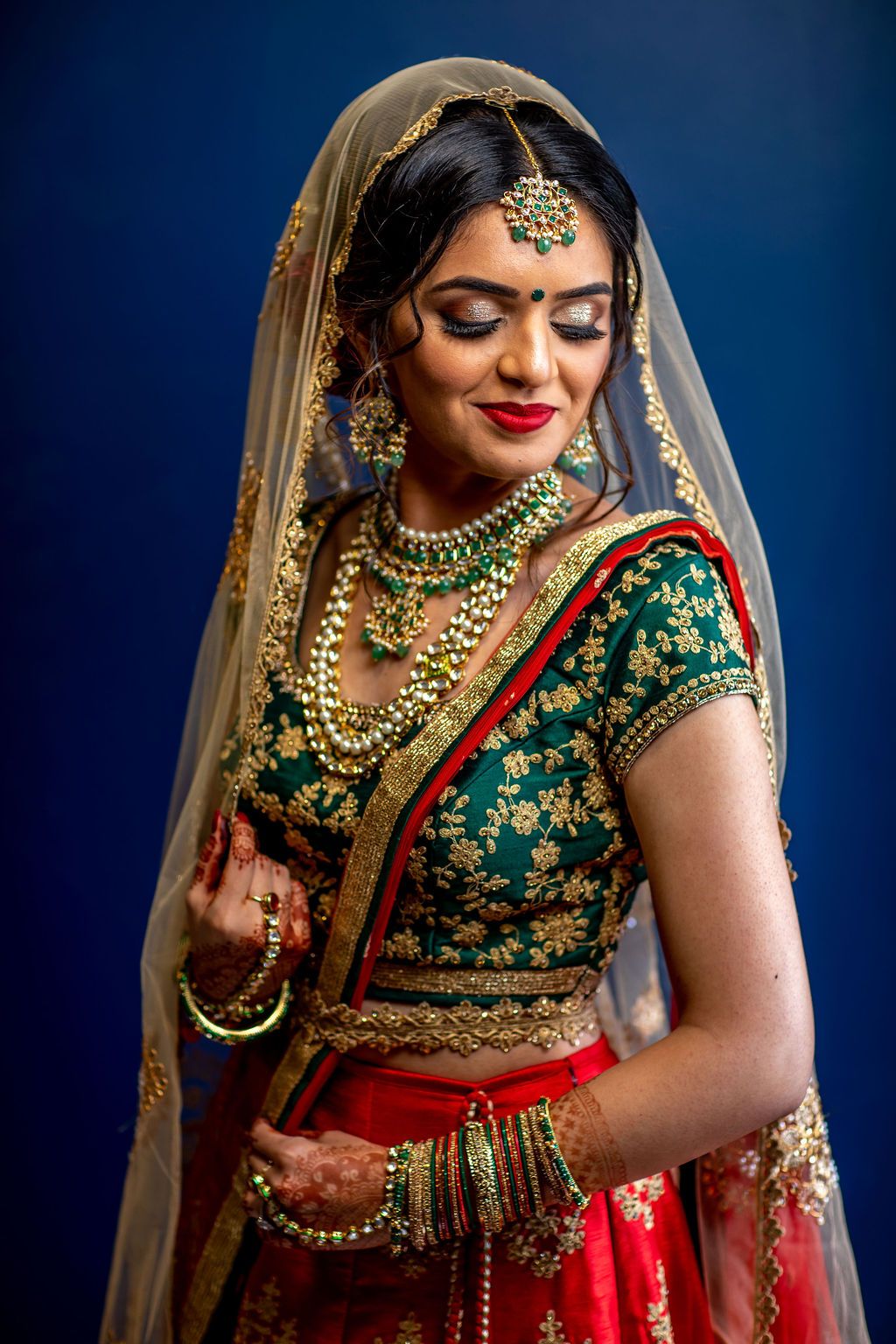 Indian bride in red and green lehenga with gold jewelry and veil poses elegantly against a blue background.