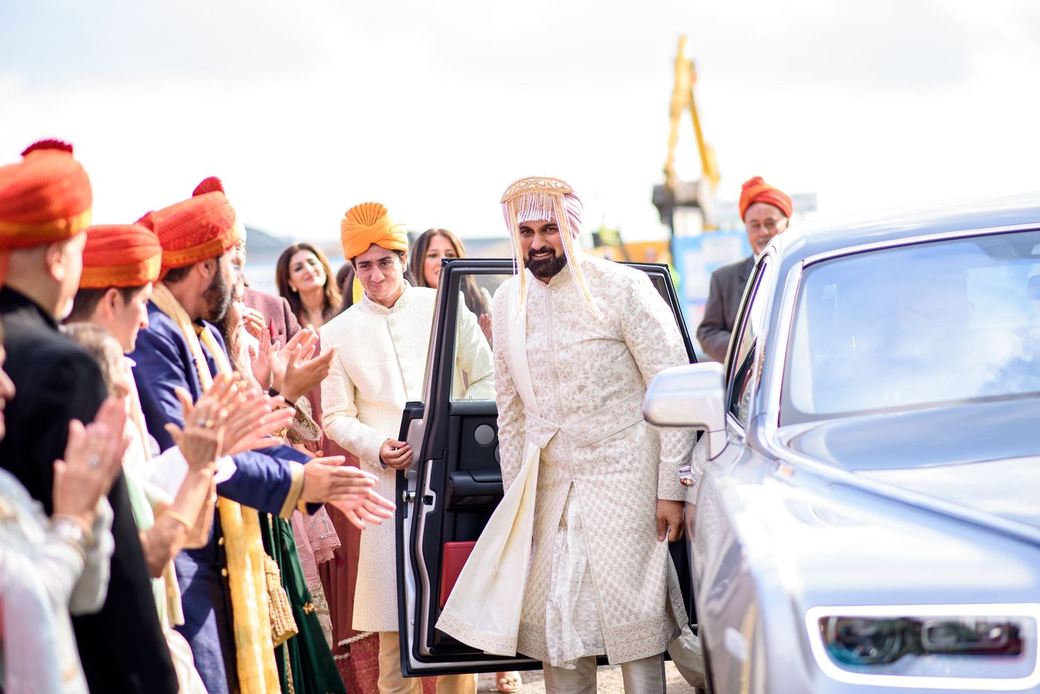 Groom in white sherwani exits luxury car greeted by guests in colorful turbans at Indian wedding ceremony.