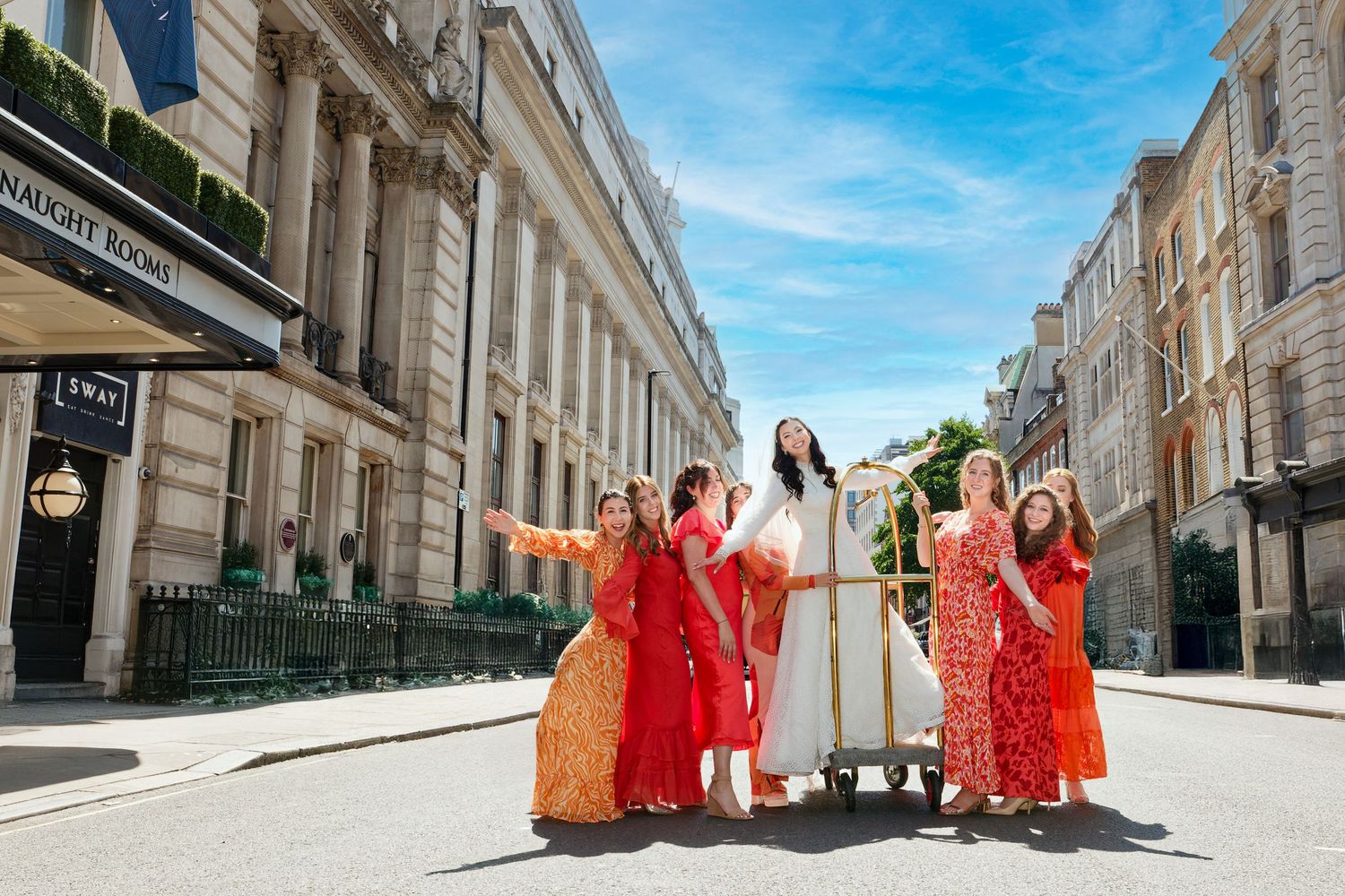 A group in bright red dresses walks down a historic European street with white stone buildings on a sunny day.