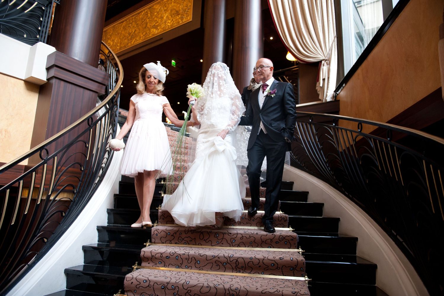 Elegant wedding party descending grand staircase in formal attire at luxurious venue.