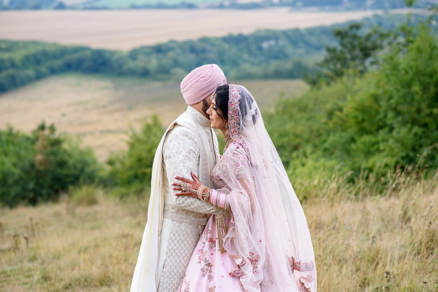 A couple in traditional South Asian wedding attire embrace on a hilltop with rolling countryside in the background.