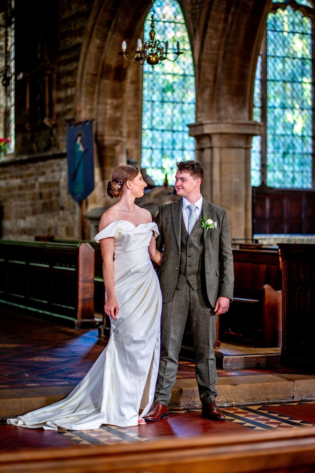 Bride and groom standing together in a historic stone church aisle, with stained glass windows and wooden pews in the background.