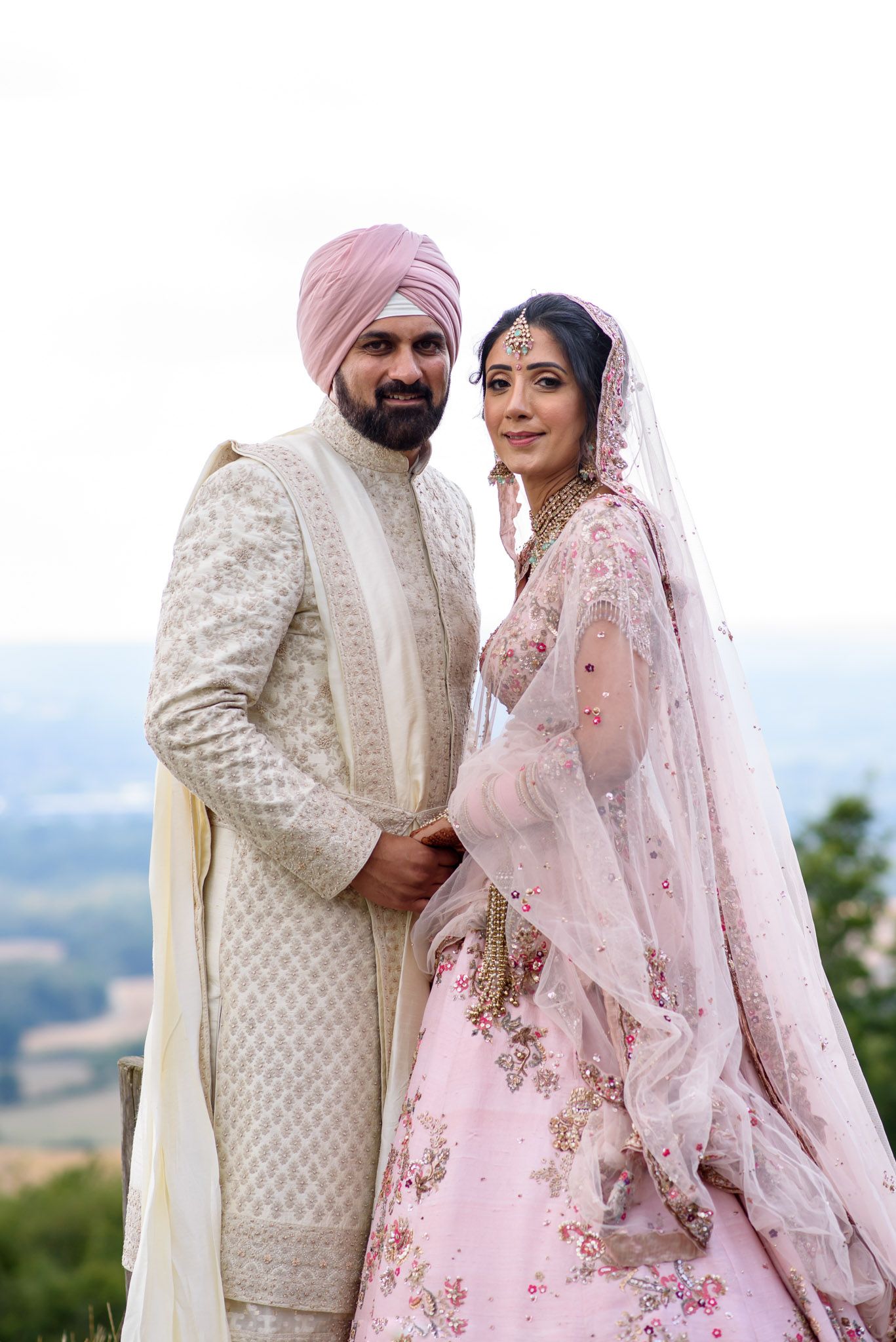 Indian couple in traditional wedding attire, groom in cream sherwani and pink turban, bride in floral pink lehenga.