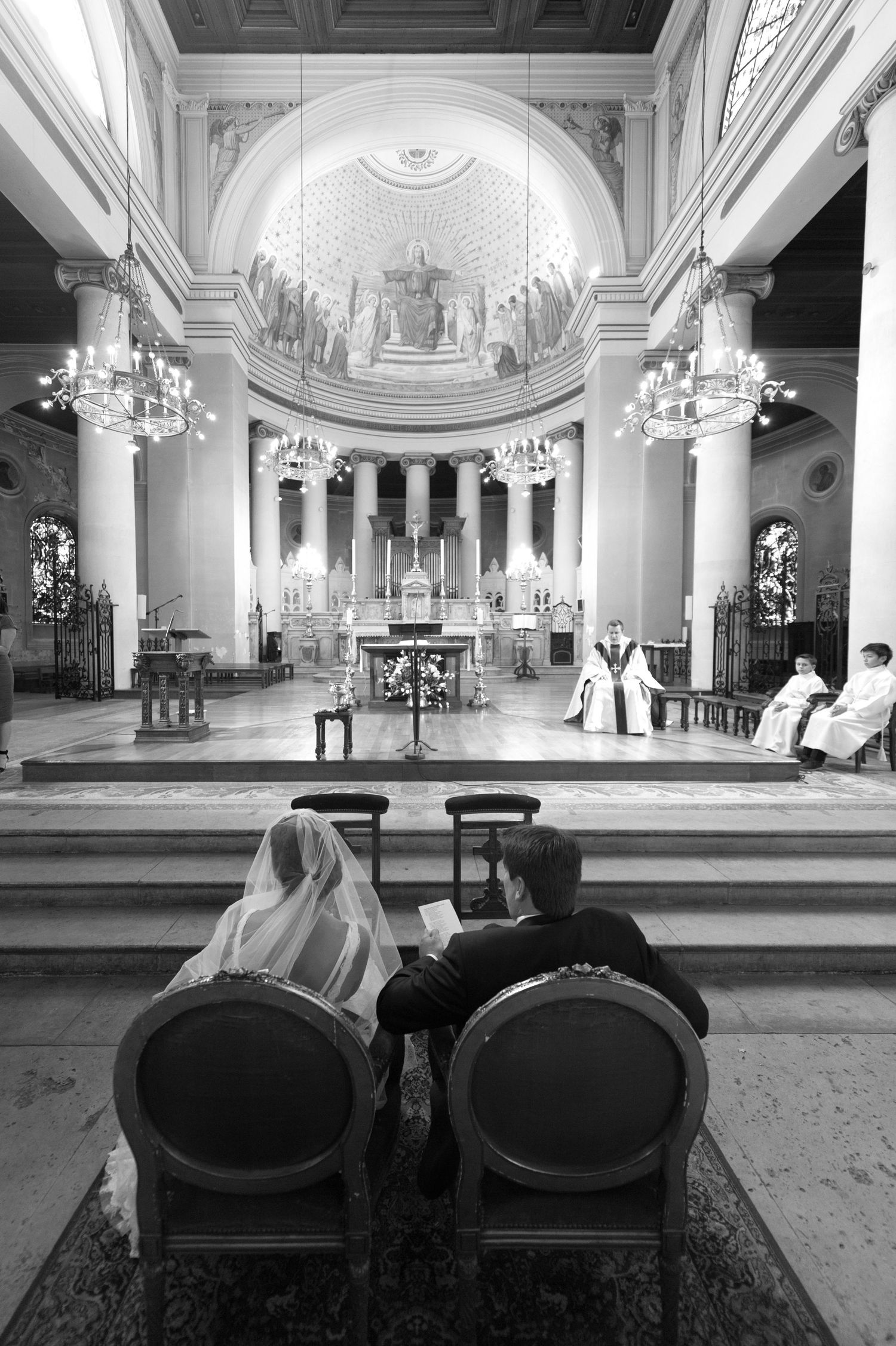 Black and white photo of a bride and groom seated in a grand church during their wedding ceremony.