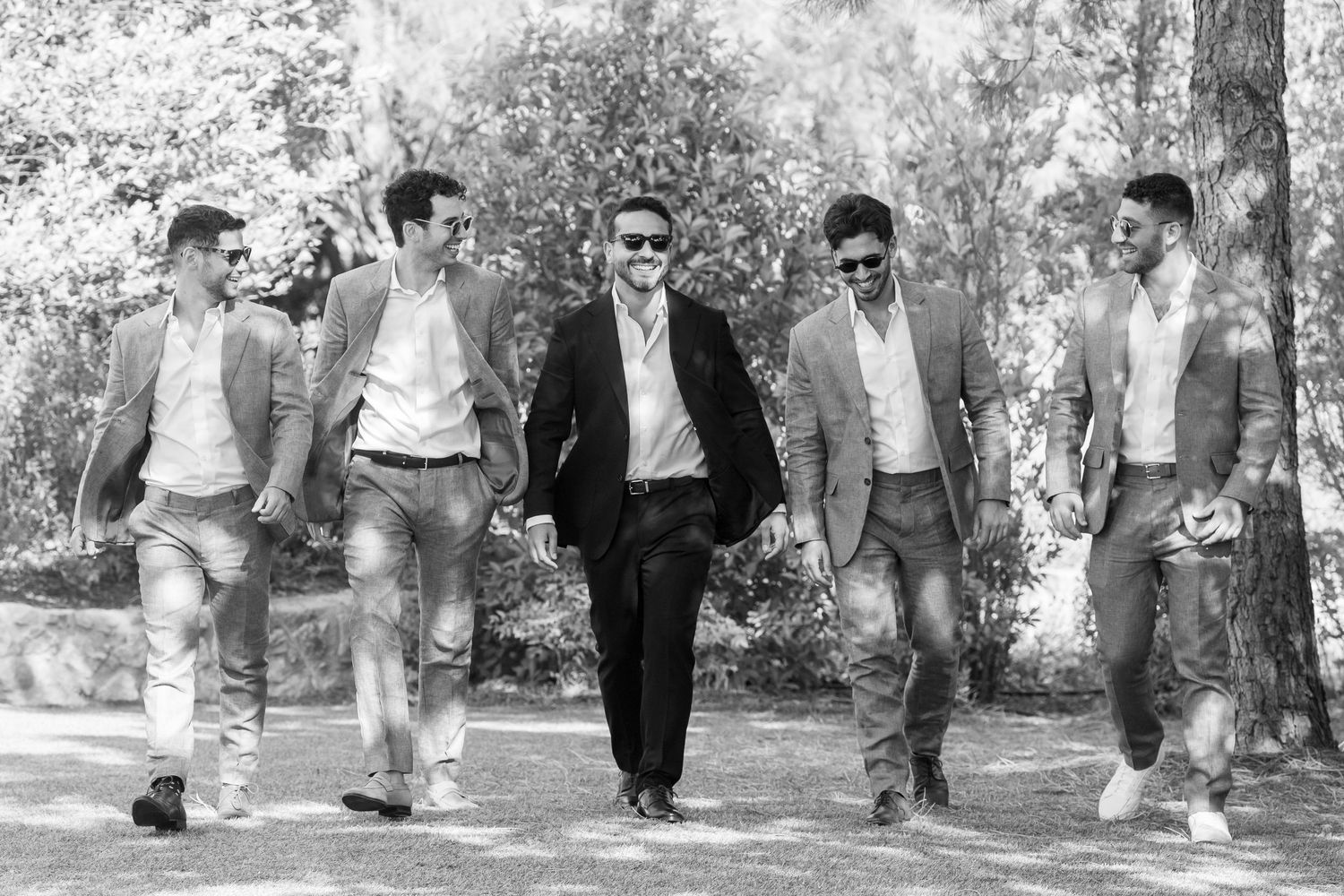 Five stylish men in suits and jeans walk confidently through a tree-lined path in a black and white photo.