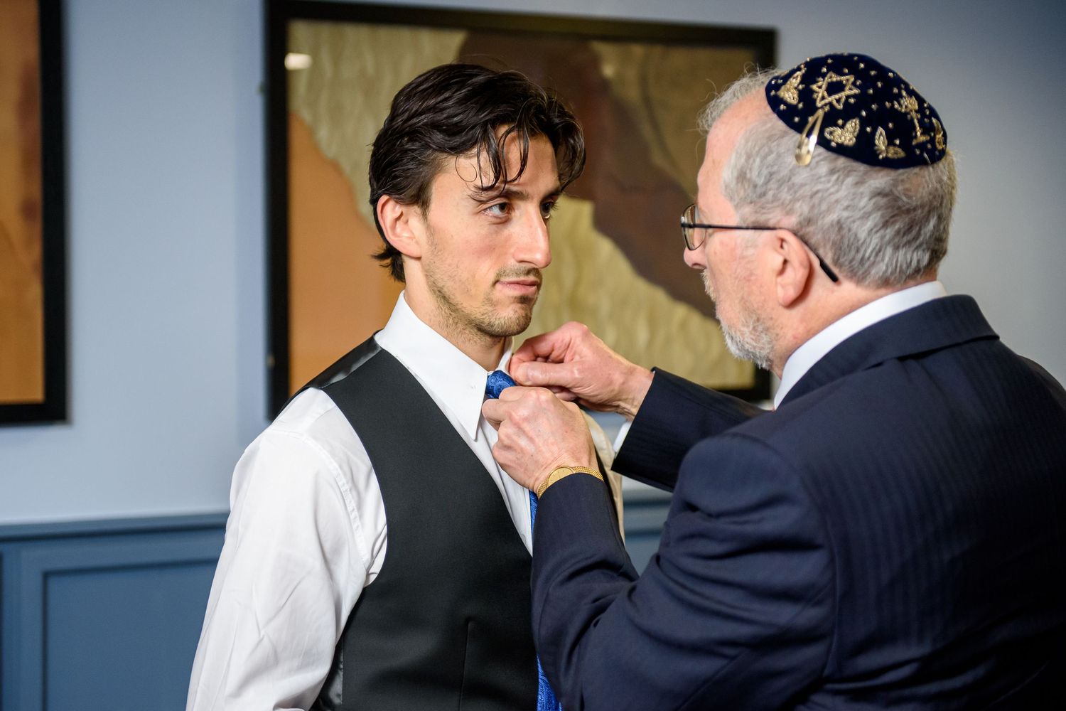 An older man wearing a yarmulke adjusts the tie of a younger man in a white shirt and black vest.