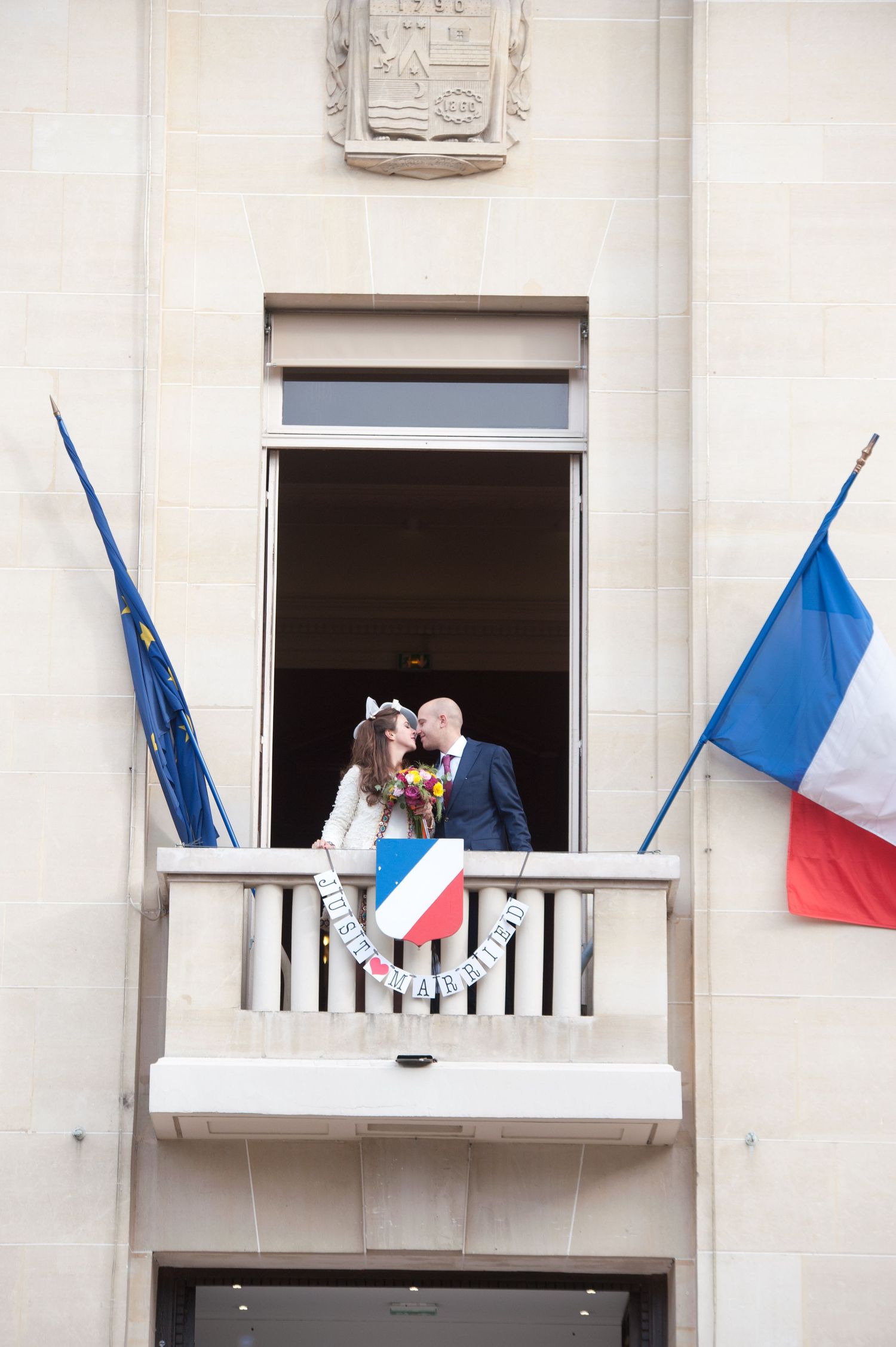 French flags wave from white balconies on a light-colored building facade.
