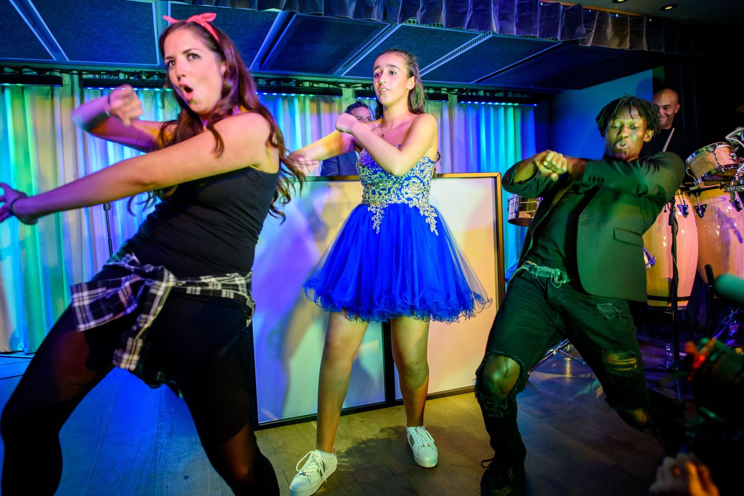 Dancers in colorful outfits perform energetic dance moves on a stage illuminated with blue and green lighting.