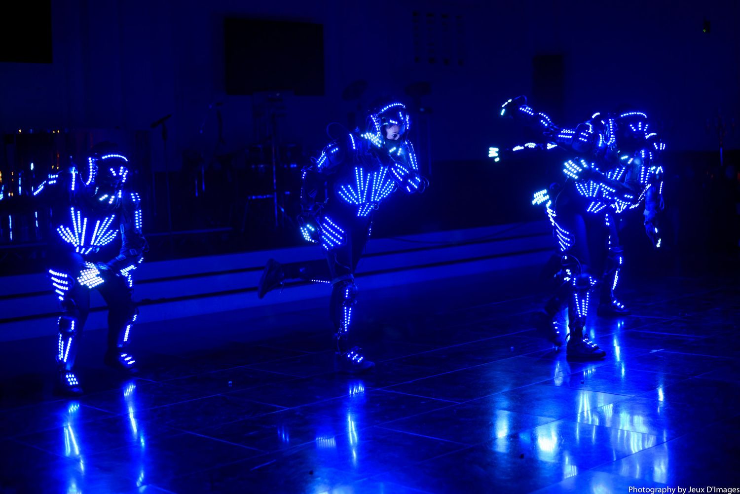 Performers in glowing LED suits dance across a dark stage creating mesmerizing blue light trails during a show.