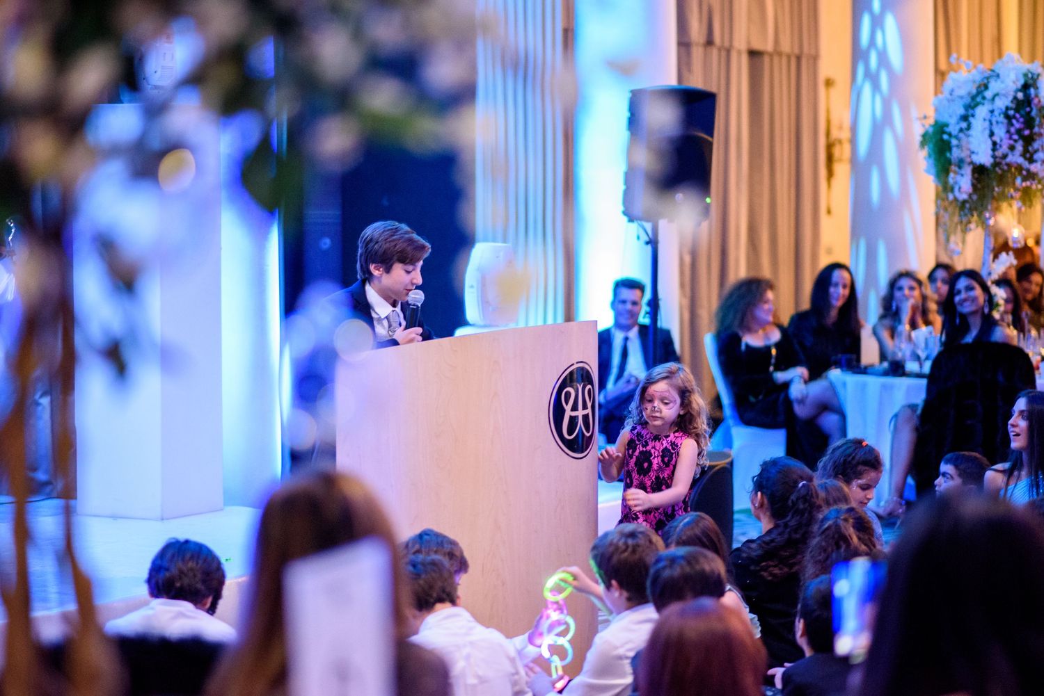 Wedding guests mingle and dance in an elegant ballroom with ornate columns and warm lighting during an evening reception.