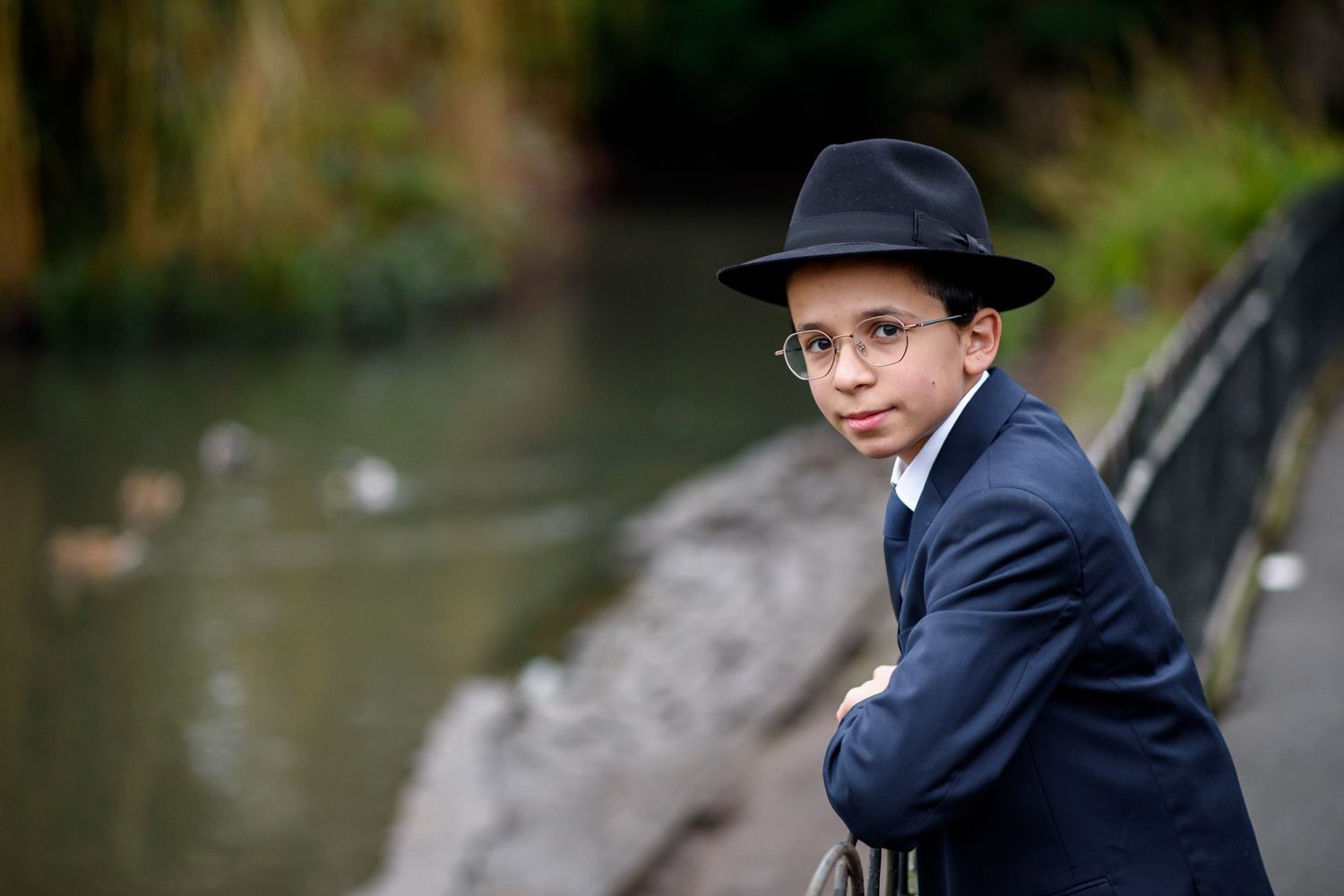 A person wearing a black hat and suit stands in a contemplative pose against a blurred outdoor background.