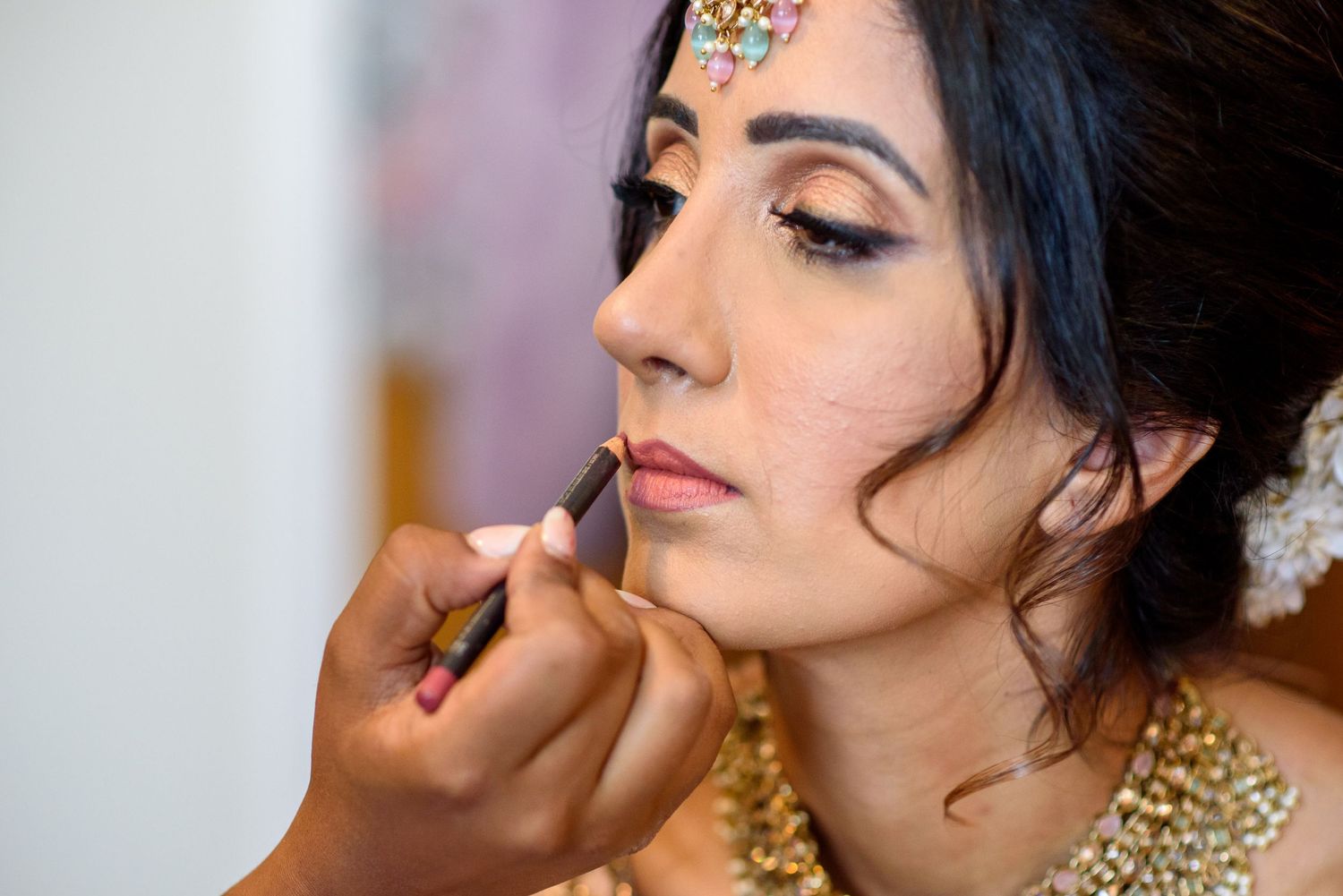 Professional makeup artist applying lipstick to client wearing traditional gold jewelry and formal attire.