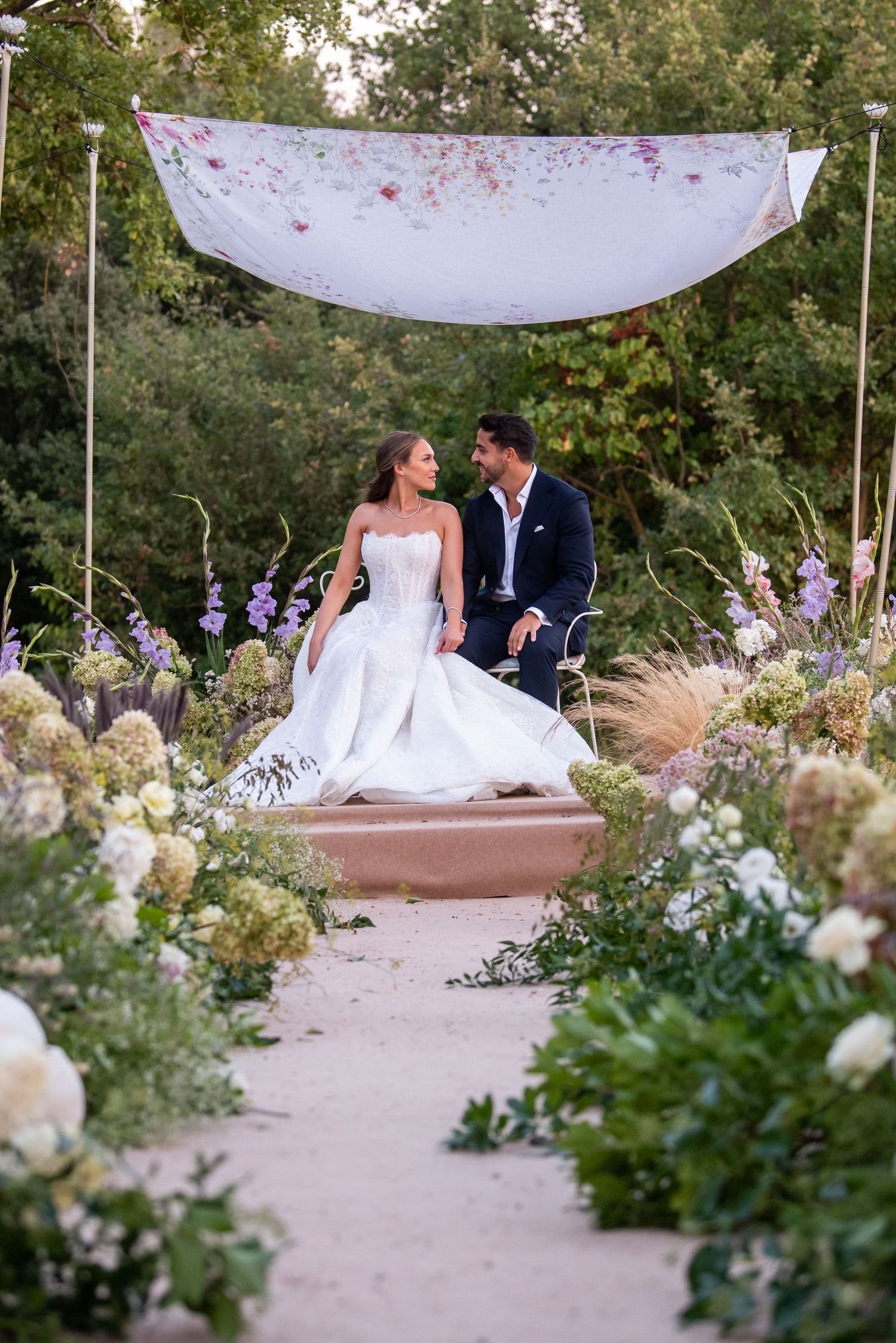 A romantic garden wedding scene with a couple sitting beneath a white canopy surrounded by lush floral arrangements.