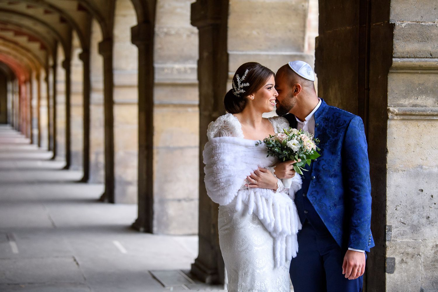 A bride and groom share a romantic moment under stone arches, she in a white gown with fur wrap, he in a blue suit with kippah.
