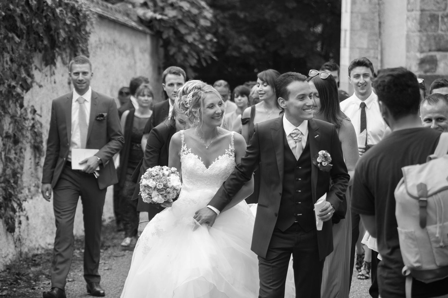 Black and white wedding photo of smiling bride and groom walking with guests outdoors near stone wall.