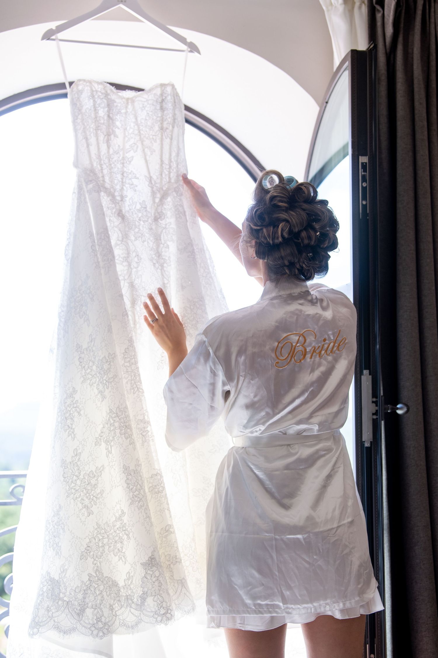 A white silk robe-clad figure admires a delicate lace wedding dress hanging by a sunlit window.