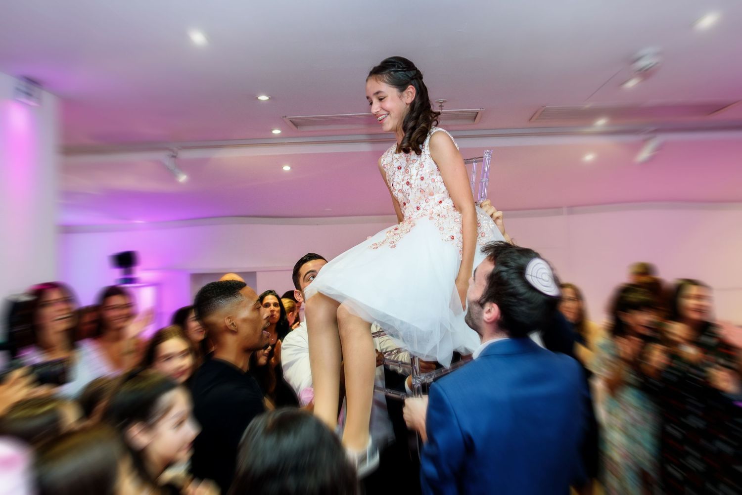 Wedding guests dance and celebrate on a white dance floor under purple mood lighting at an indoor reception venue.