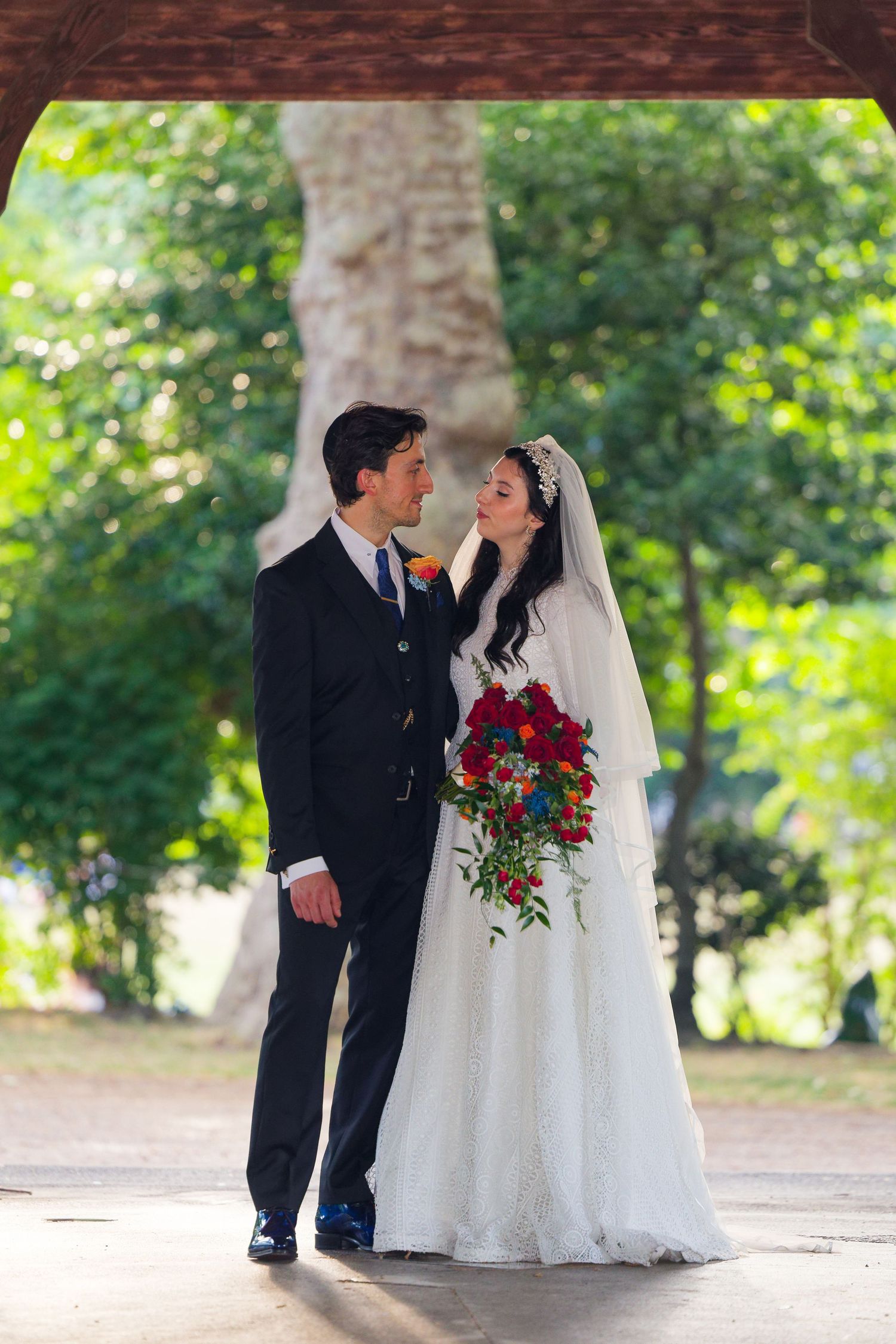A romantic wedding couple in formal attire poses together outdoors in a garden setting with lush greenery.