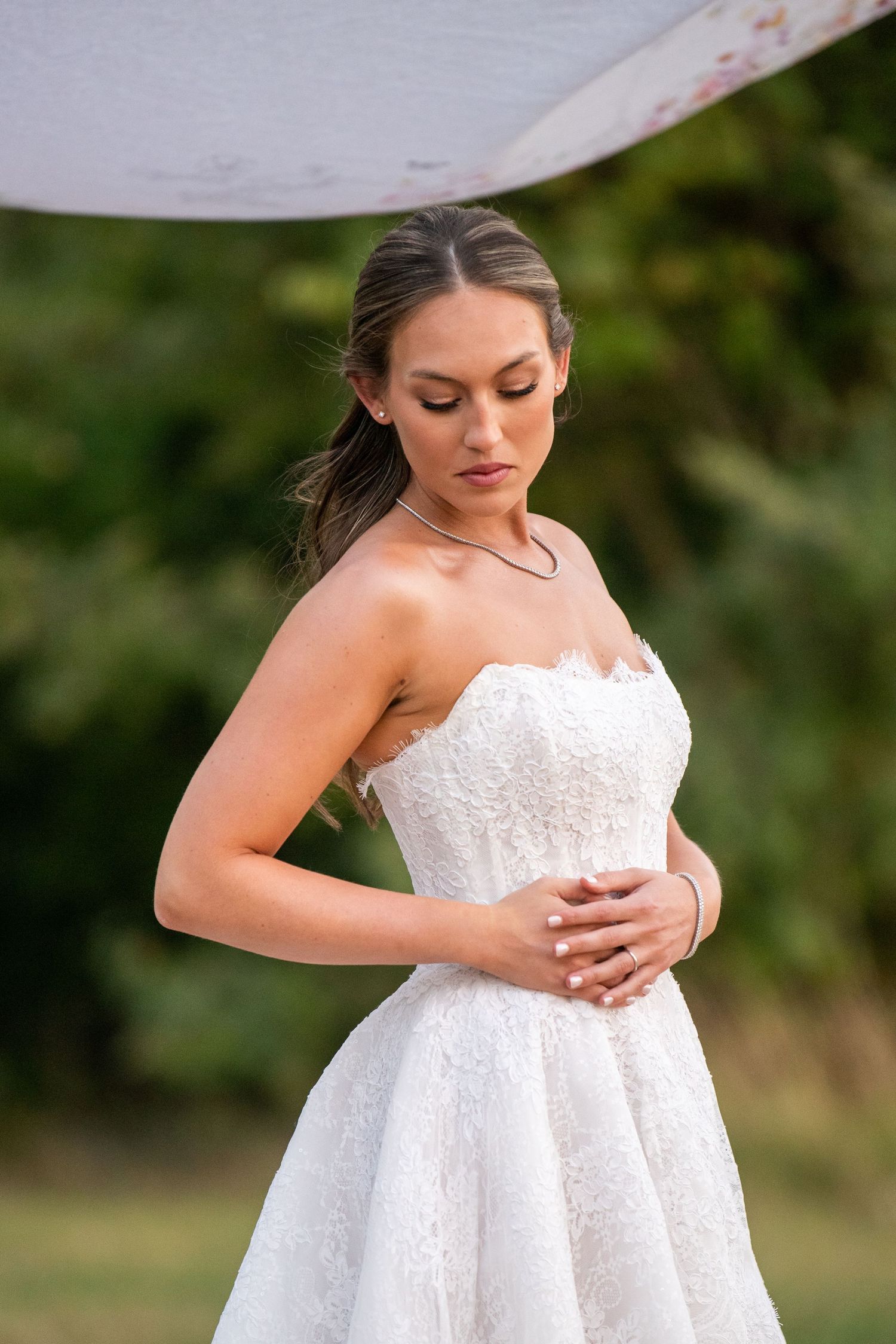 A bride in a strapless white lace wedding dress stands outdoors with a soft, natural background.