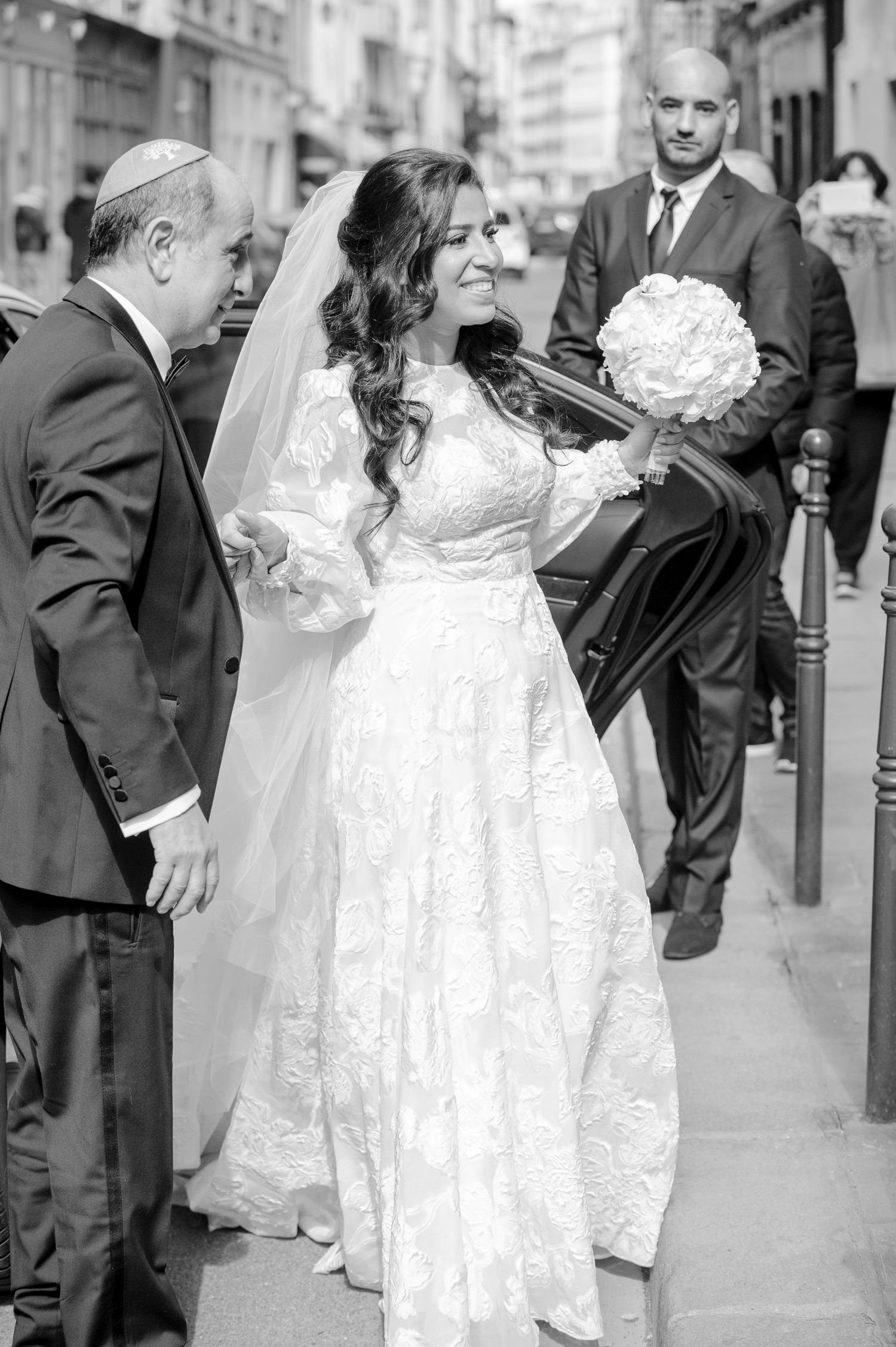 Black and white wedding photo series showing a bride in lace dress greeting guests on a city street.