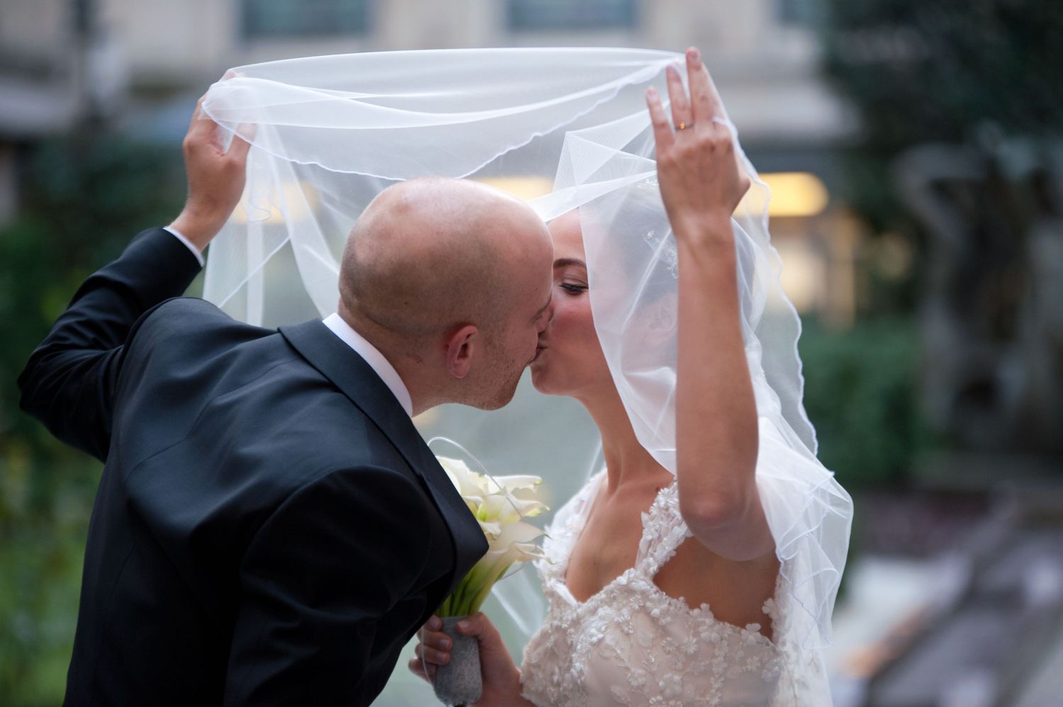 A couple in wedding attire lifts a sheer white veil together during an outdoor ceremony.