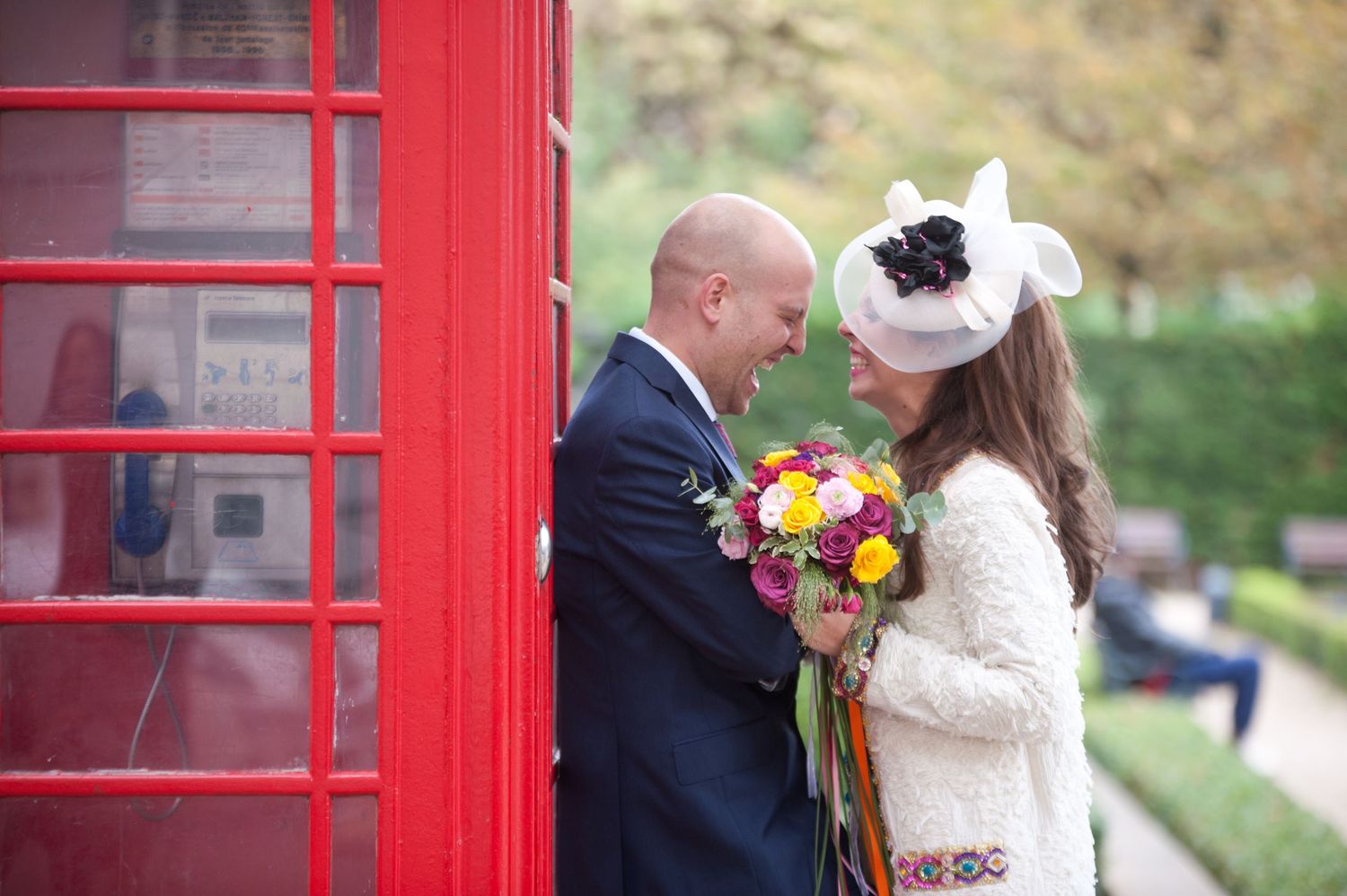 A couple embraces and kisses in front of a bright red British phone booth during a wedding photo session.
