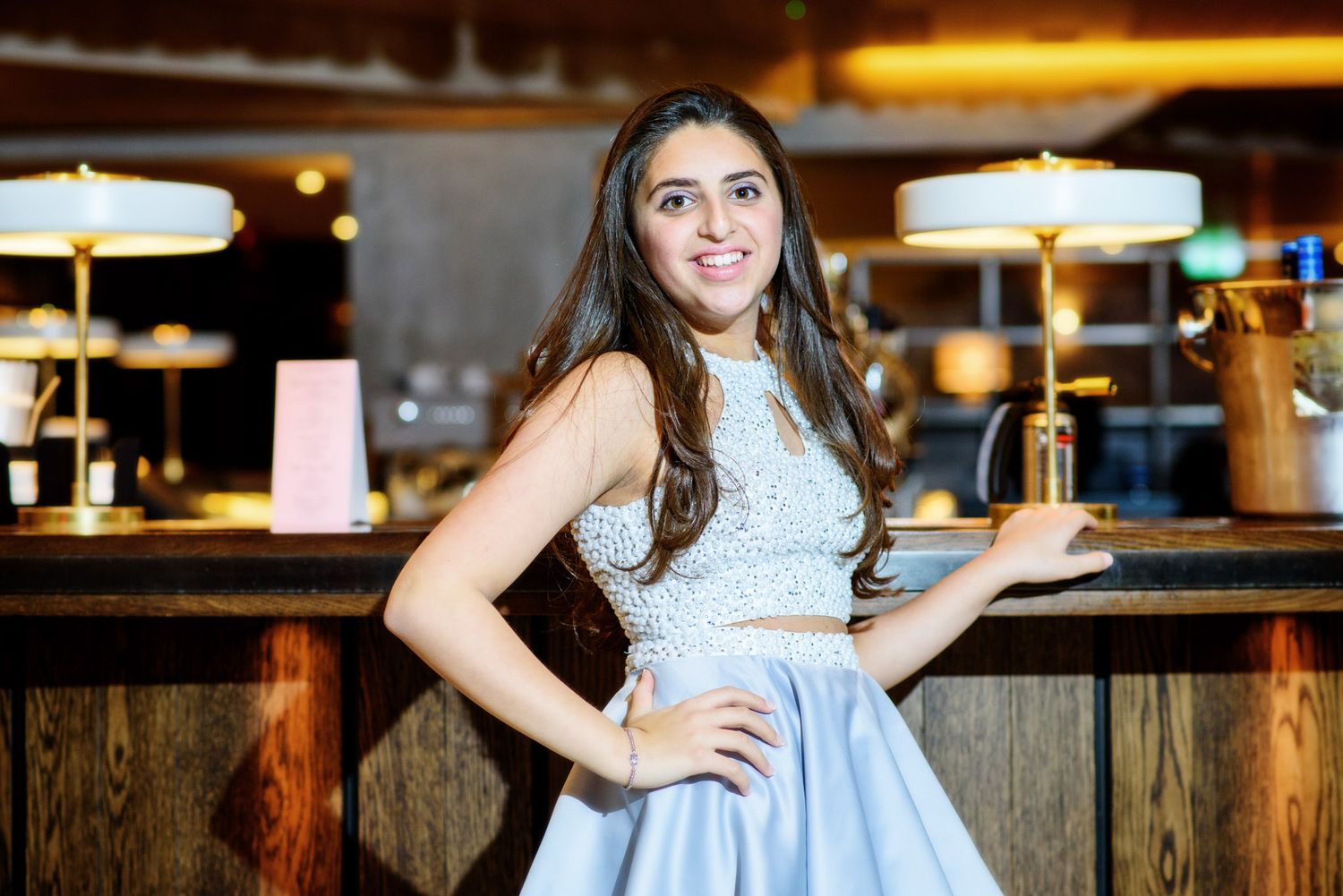 A person in a light blue dress poses near a rustic wooden fence at dusk with warm lighting in the background.