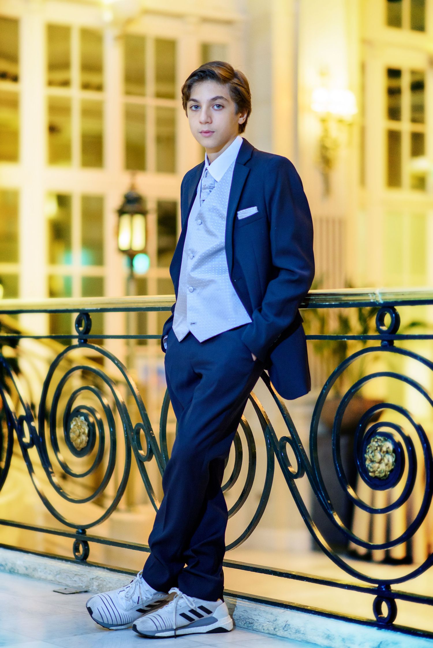 A young person in formal navy suit and white vest poses casually on ornate metal railings at an elegant venue.