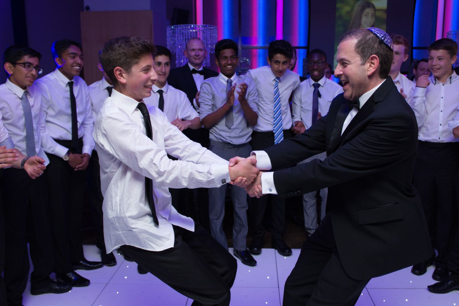 A group of performers in formal attire dancing on stage with colorful purple and blue lighting in the background.