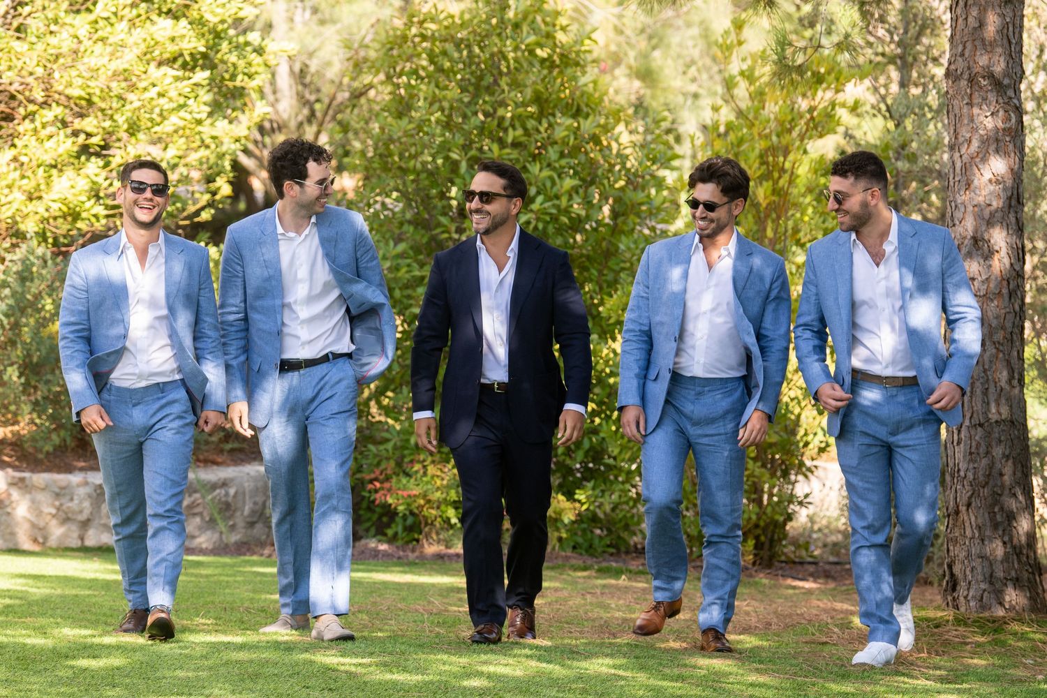 People wearing coordinated blue suits and white shirts walking in a line through a sunlit park setting.