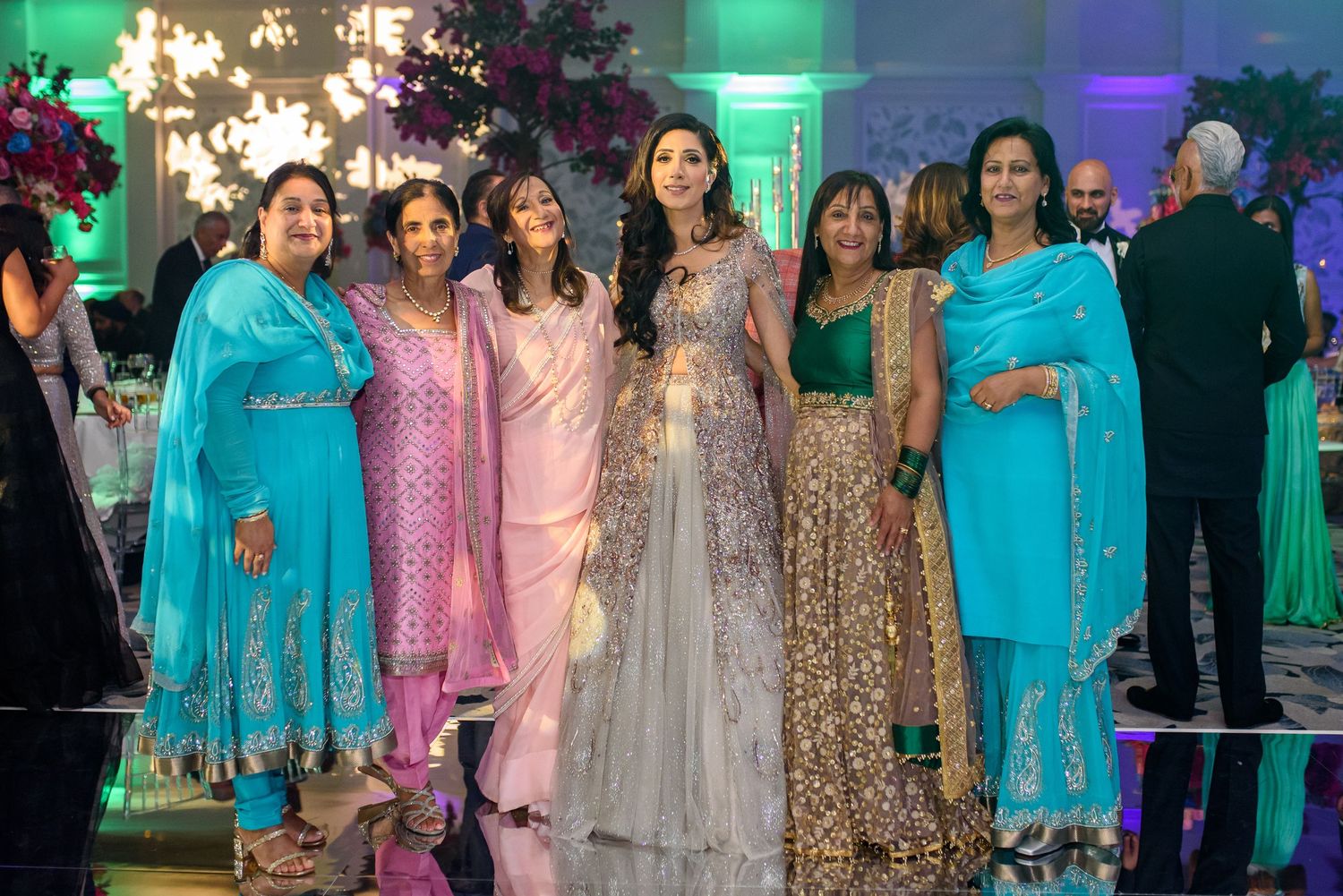 A festive celebration with people wearing traditional Indian attire in turquoise, pink and gold under colorful lighting.