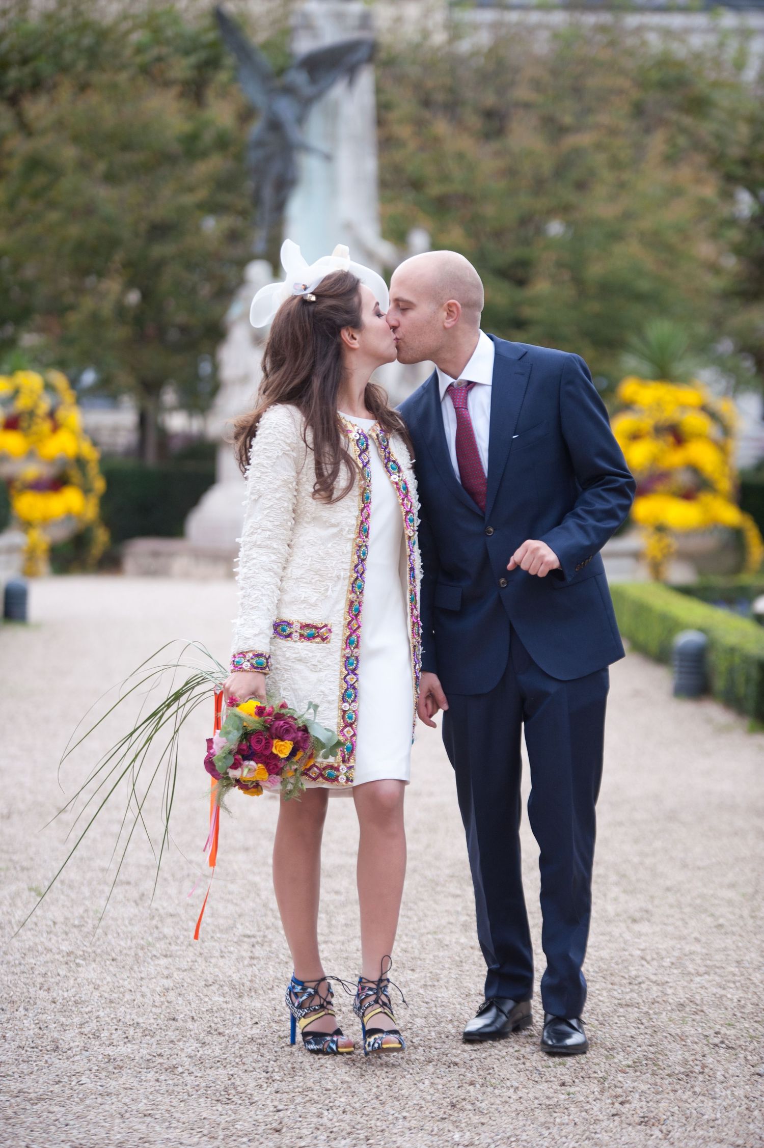 A romantic wedding couple shares a kiss on a gravel path with yellow flowers blooming in the background.