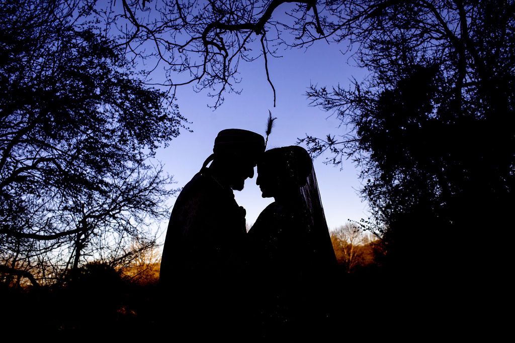 Romantic silhouette of a couple facing each other at twilight, framed by dark tree branches against a blue and orange sky.