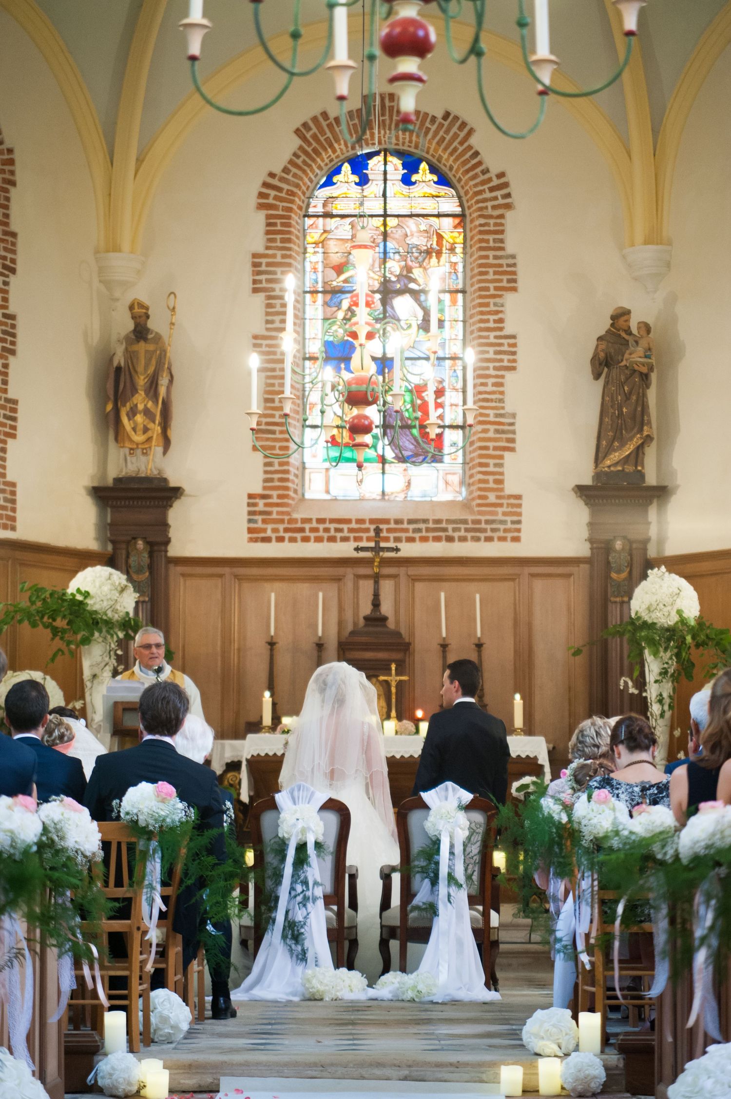 Bride and groom at church altar with stained glass window, floral arrangements, and candles during wedding ceremony.