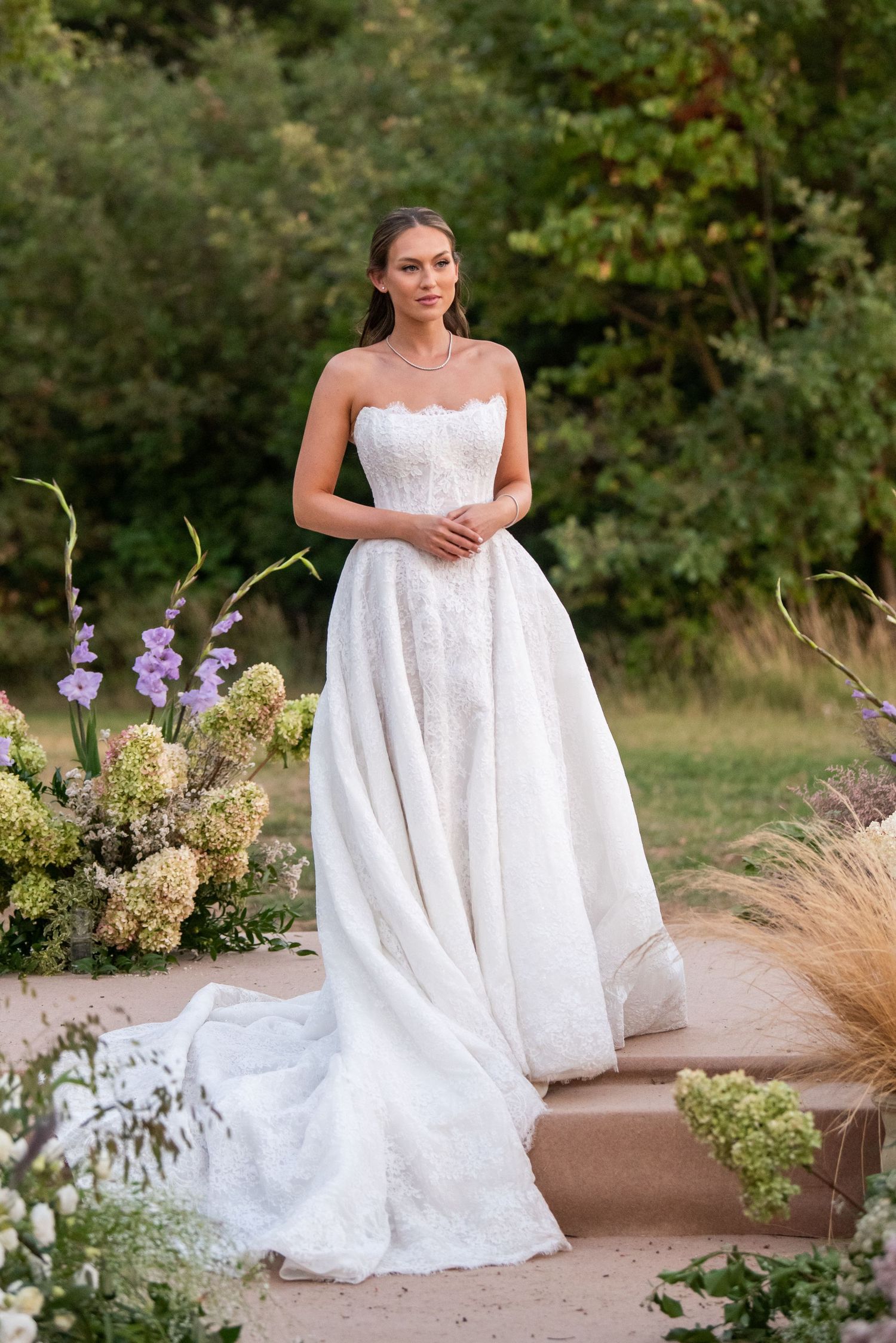 A bridal portrait session in a garden setting with white flowers and greenery showcases an elegant lace wedding gown.