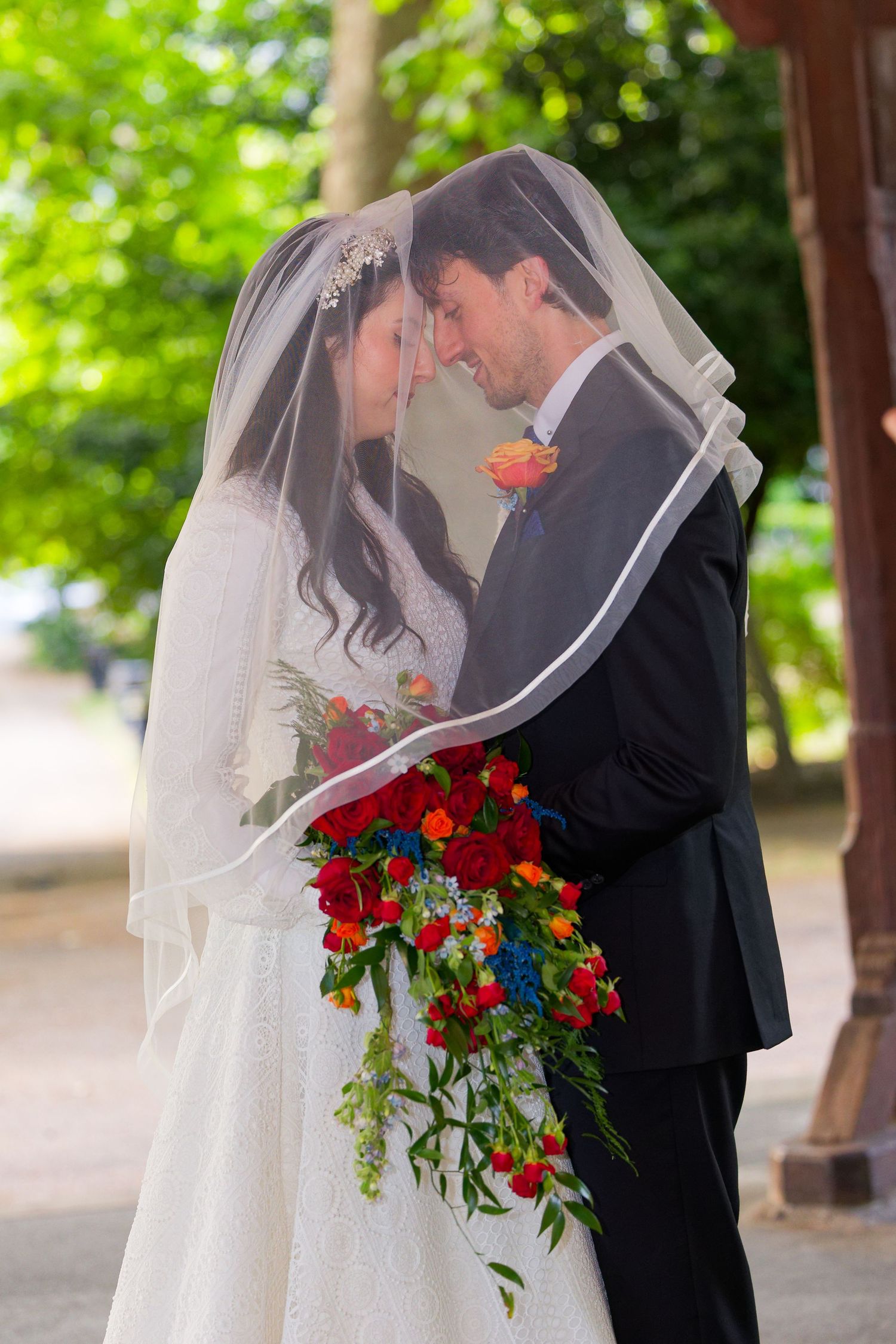 Newlywed couple sharing an intimate moment with a vibrant red rose bouquet under a tree-lined path.