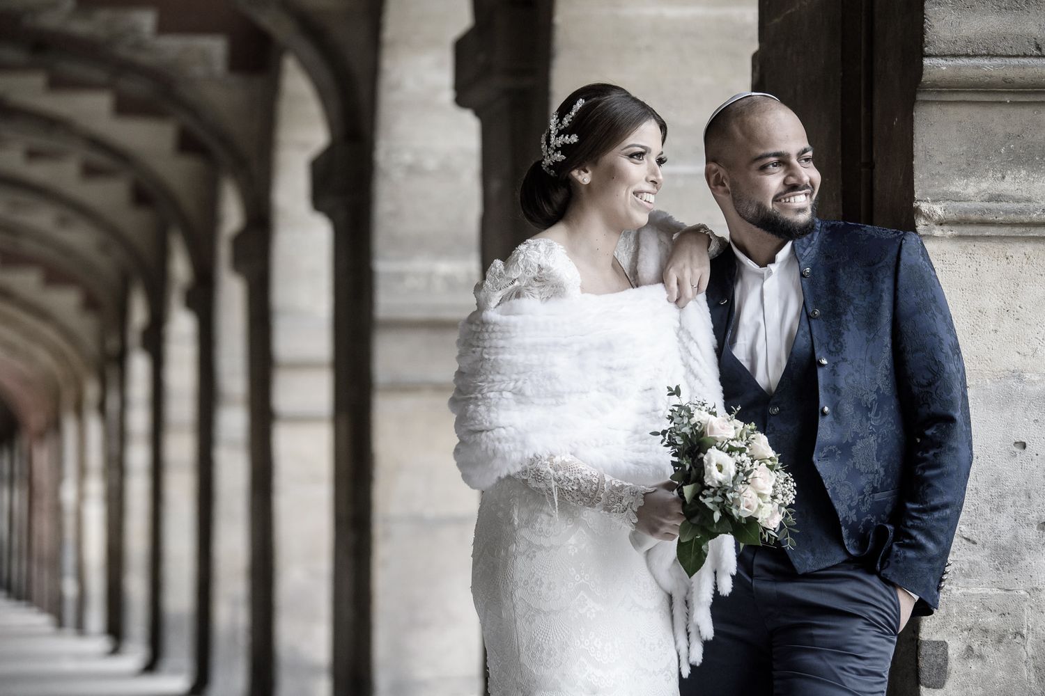 Bride in white gown and groom in blue suit share intimate moment in classical archway setting.