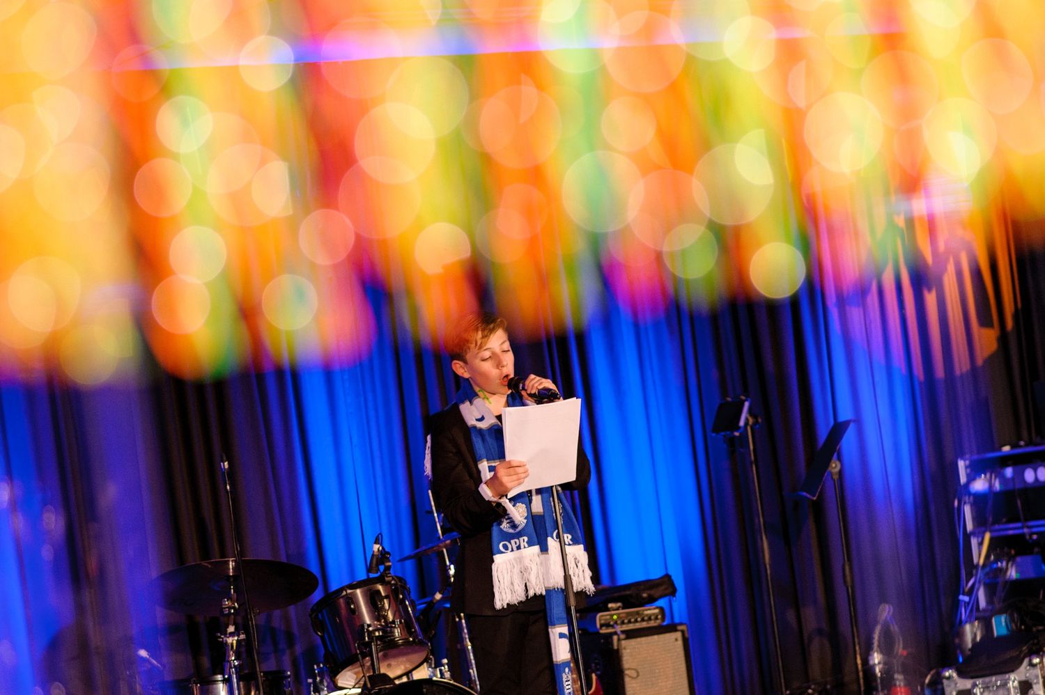 A person stands at a podium on stage with colorful rainbow lighting and illuminated vertical panels in the background.