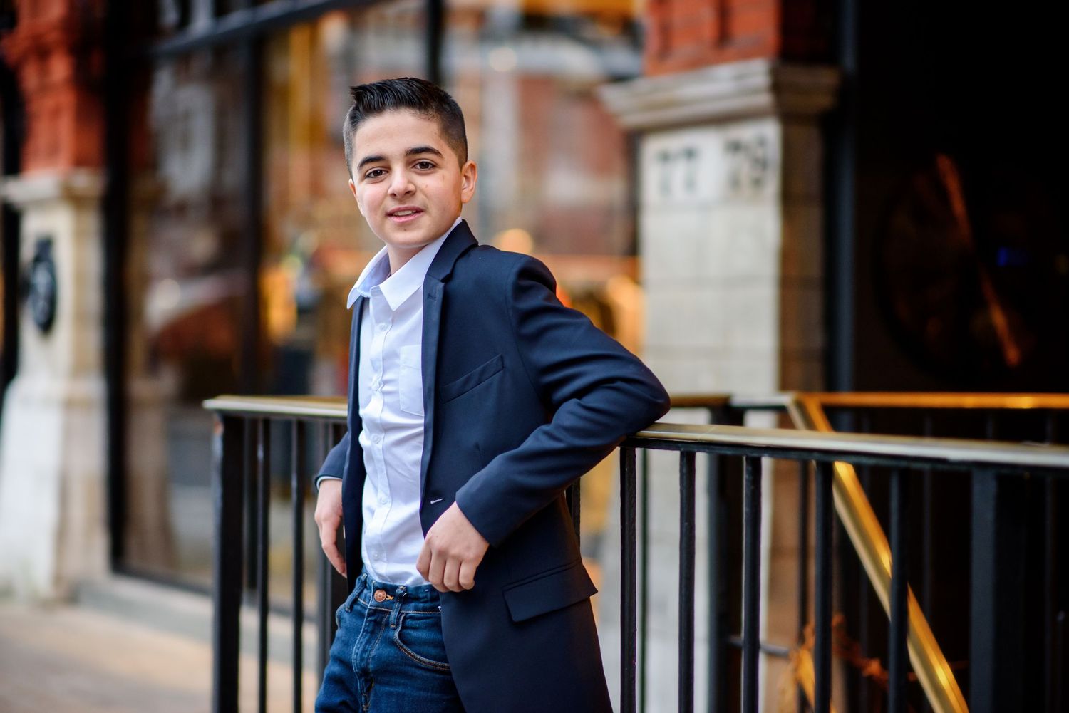 Young professional in navy blazer and jeans leans against railing outside brick building in urban setting.