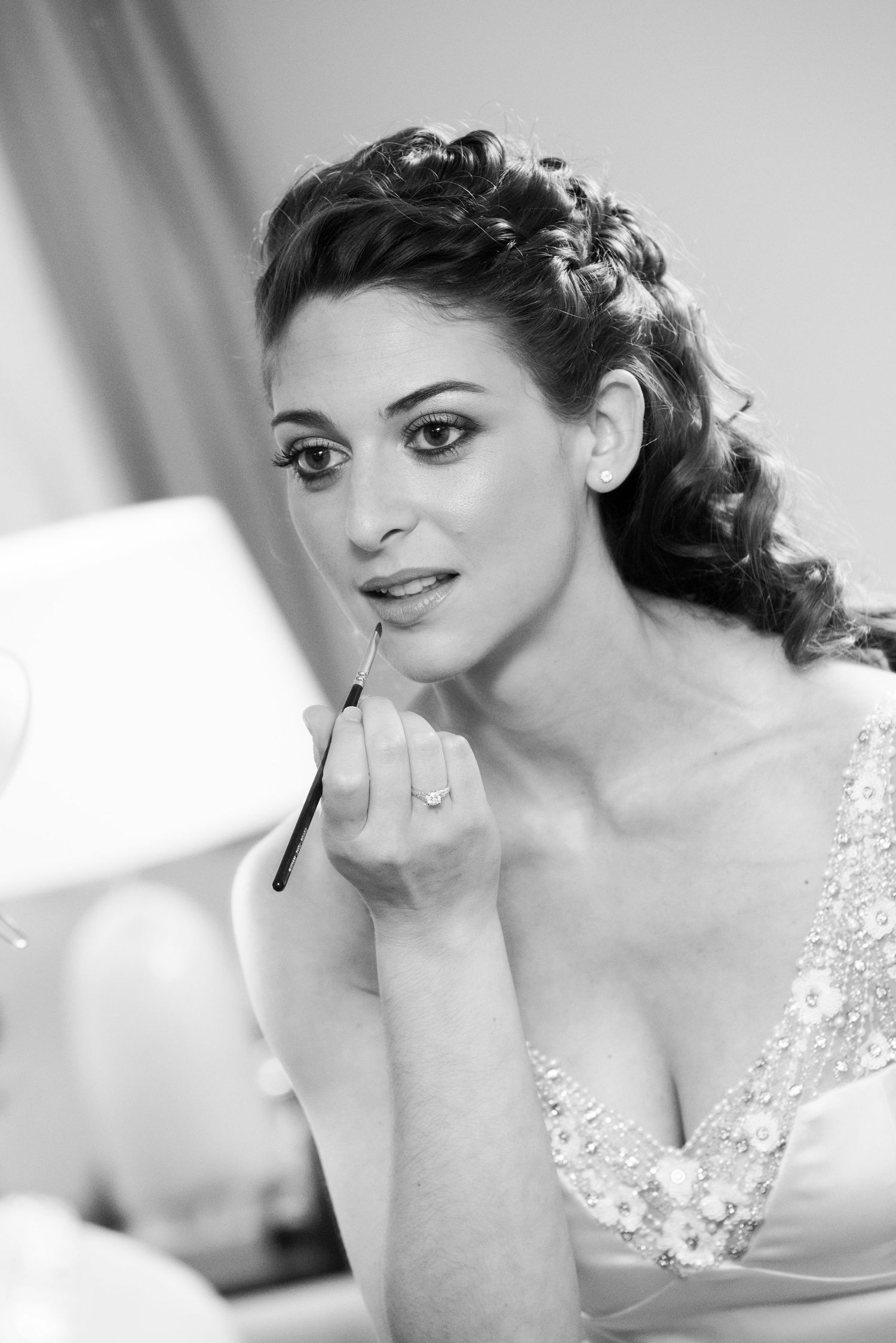 Black and white photo of a bride applying lipstick with a brush, wearing an elegant gown with floral details.