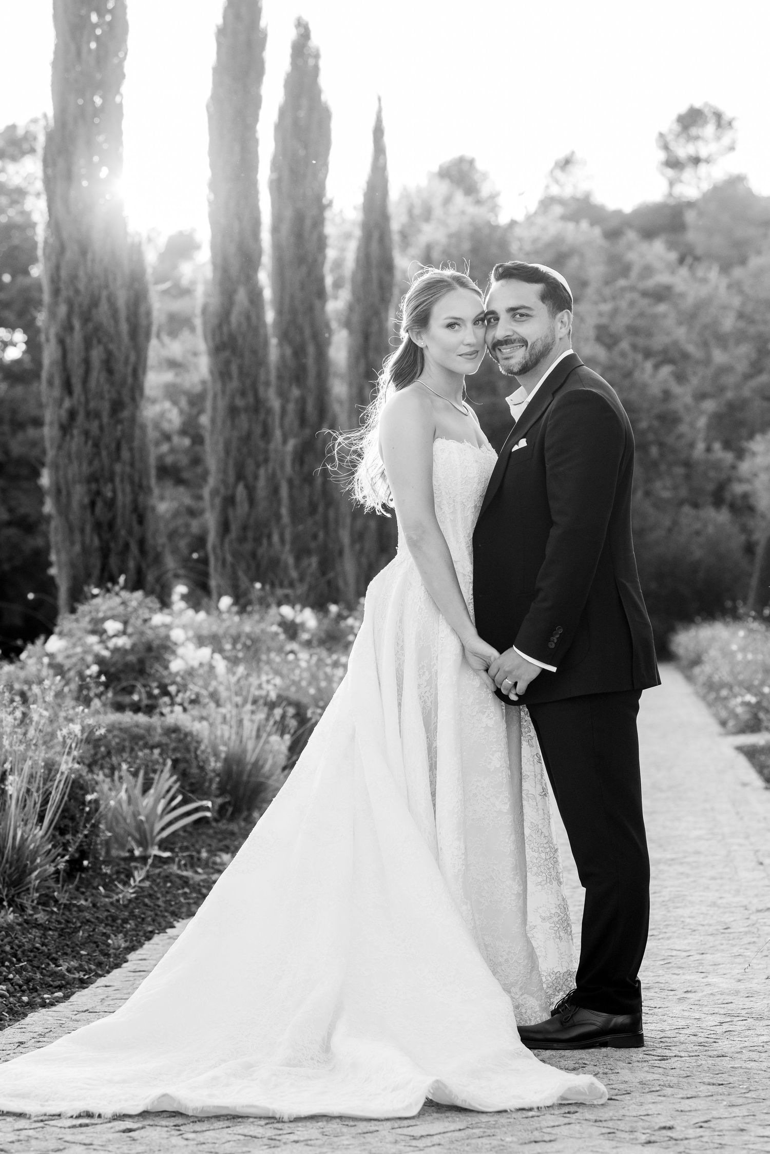 Black and white photo of a bride and groom holding hands on a garden path, surrounded by tall cypress trees.