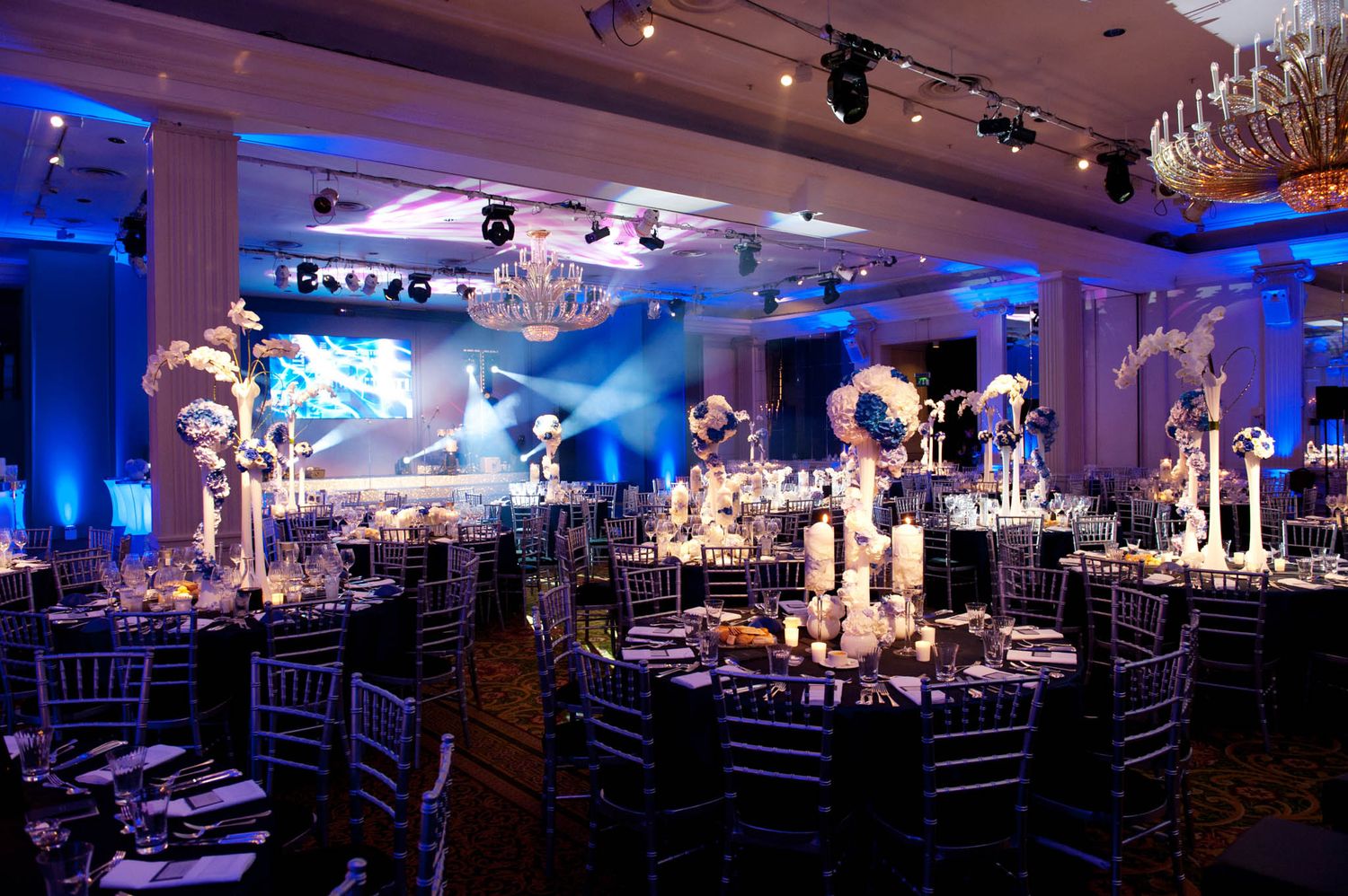 Elegant ballroom with blue uplighting features crystal chandeliers and round dining tables set for a formal event.