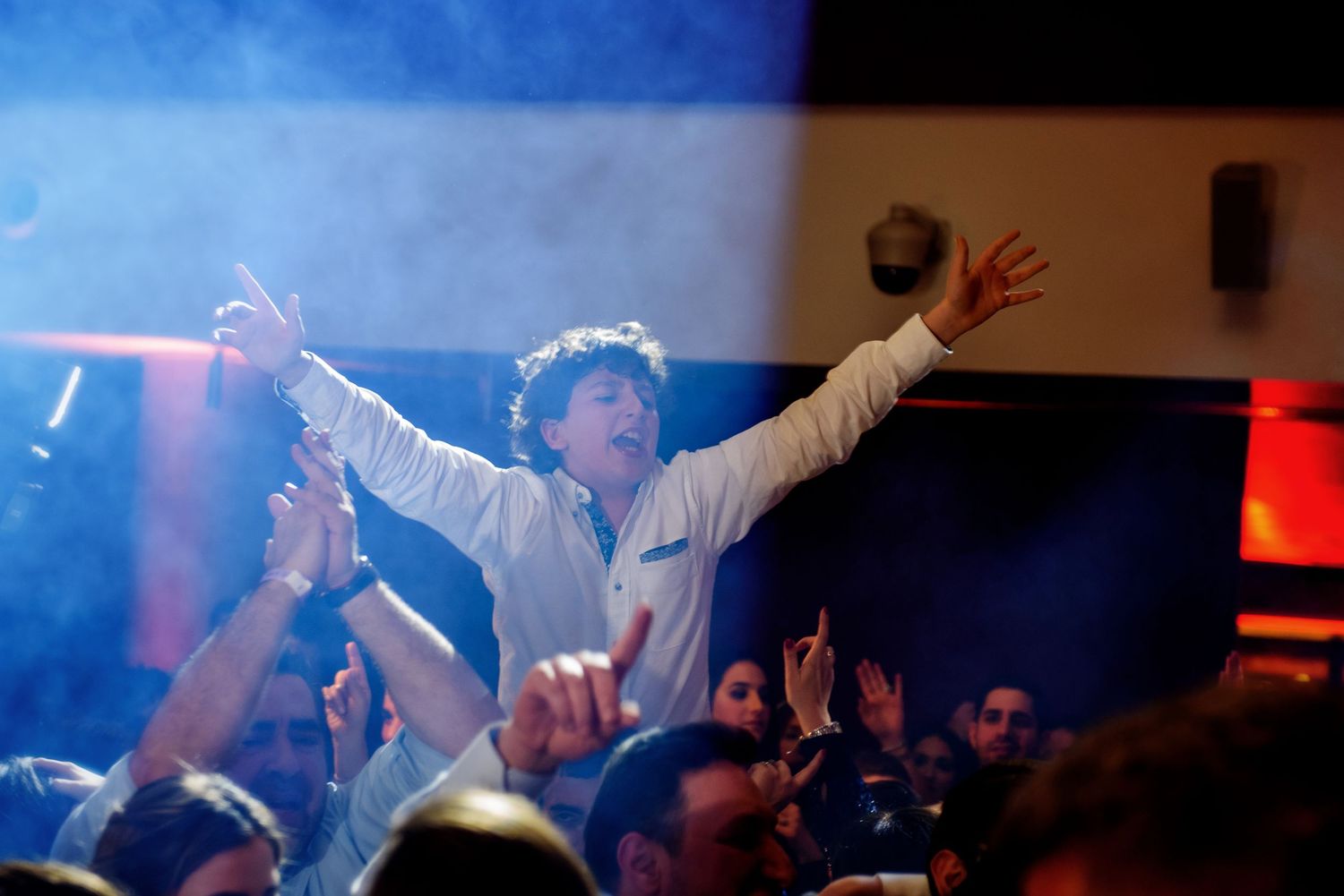 Concert crowd raises their hands in excitement during a performance with dramatic blue and red lighting.