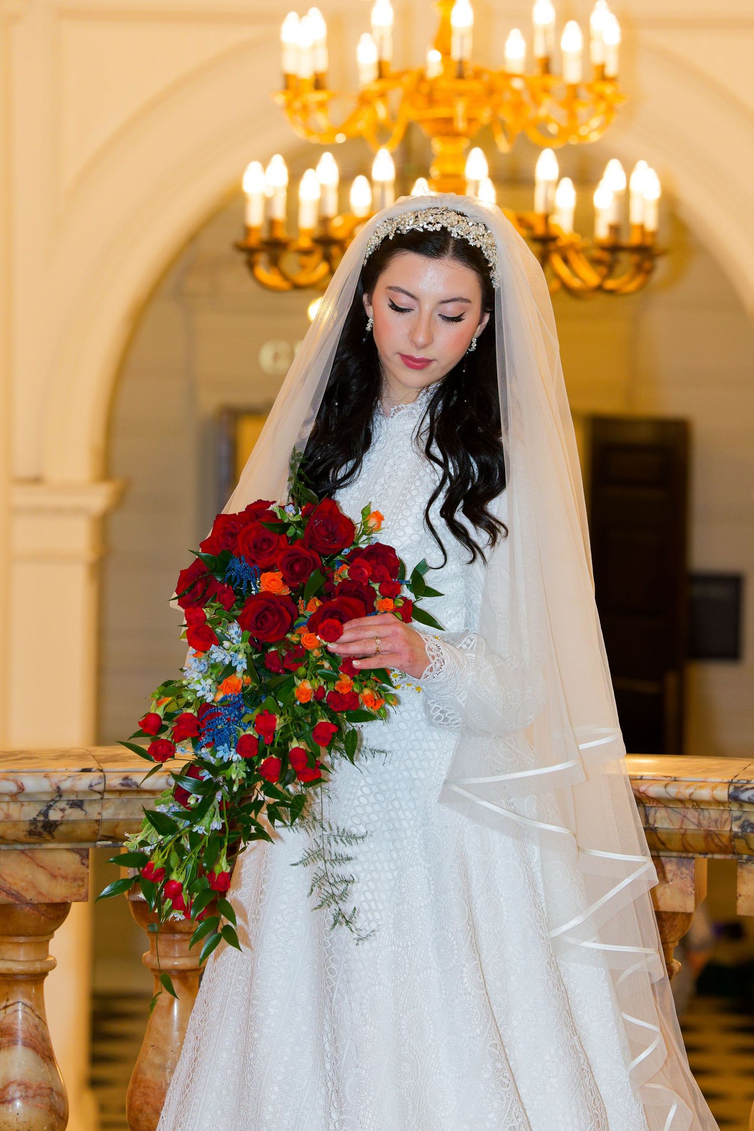 Elegant bride in white lace gown holds vibrant red and blue bouquet in luxurious venue with crystal chandeliers.