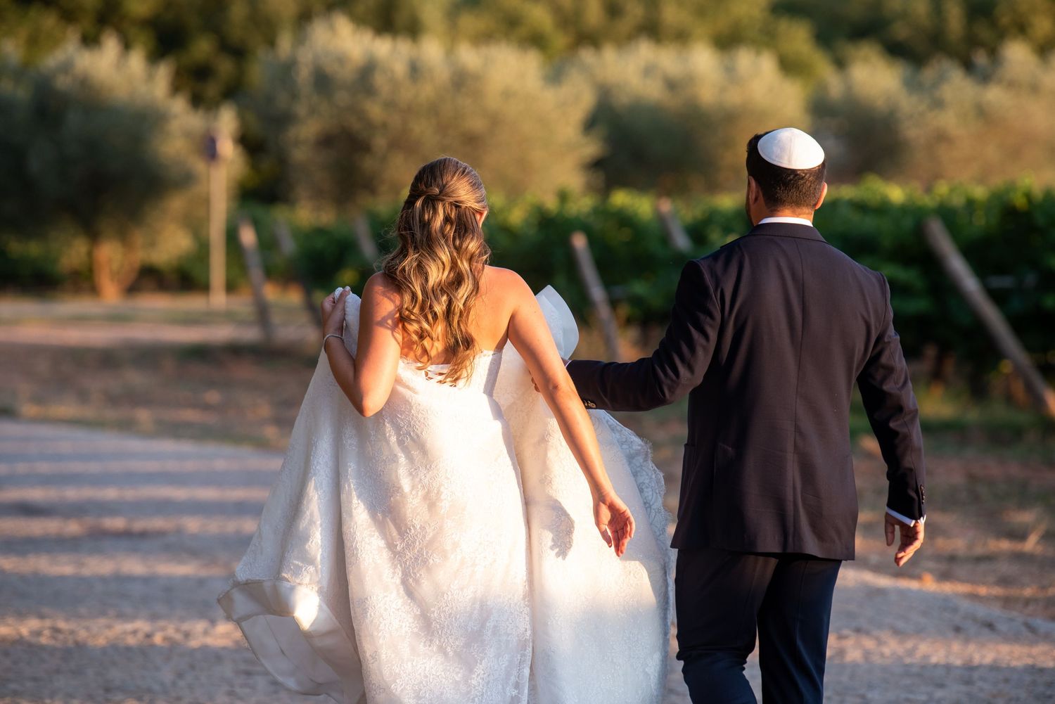 A couple in wedding attire walking away from the camera during golden hour on a sunlit path.