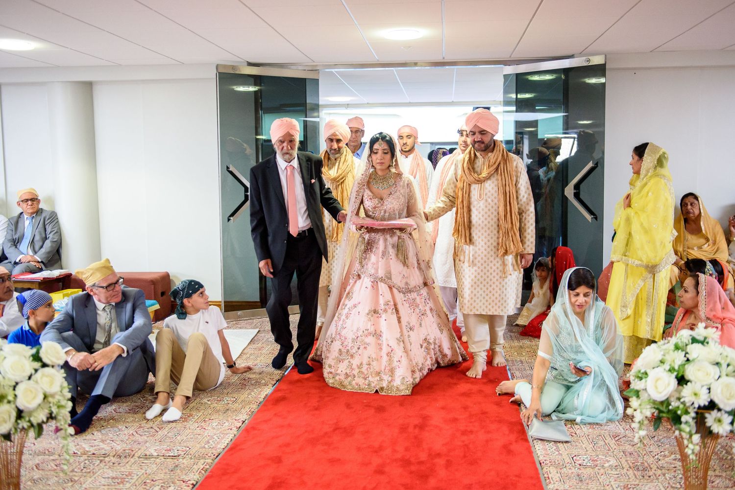 Wedding guests walk through an elegant hallway decorated with floral arrangements and red carpet.