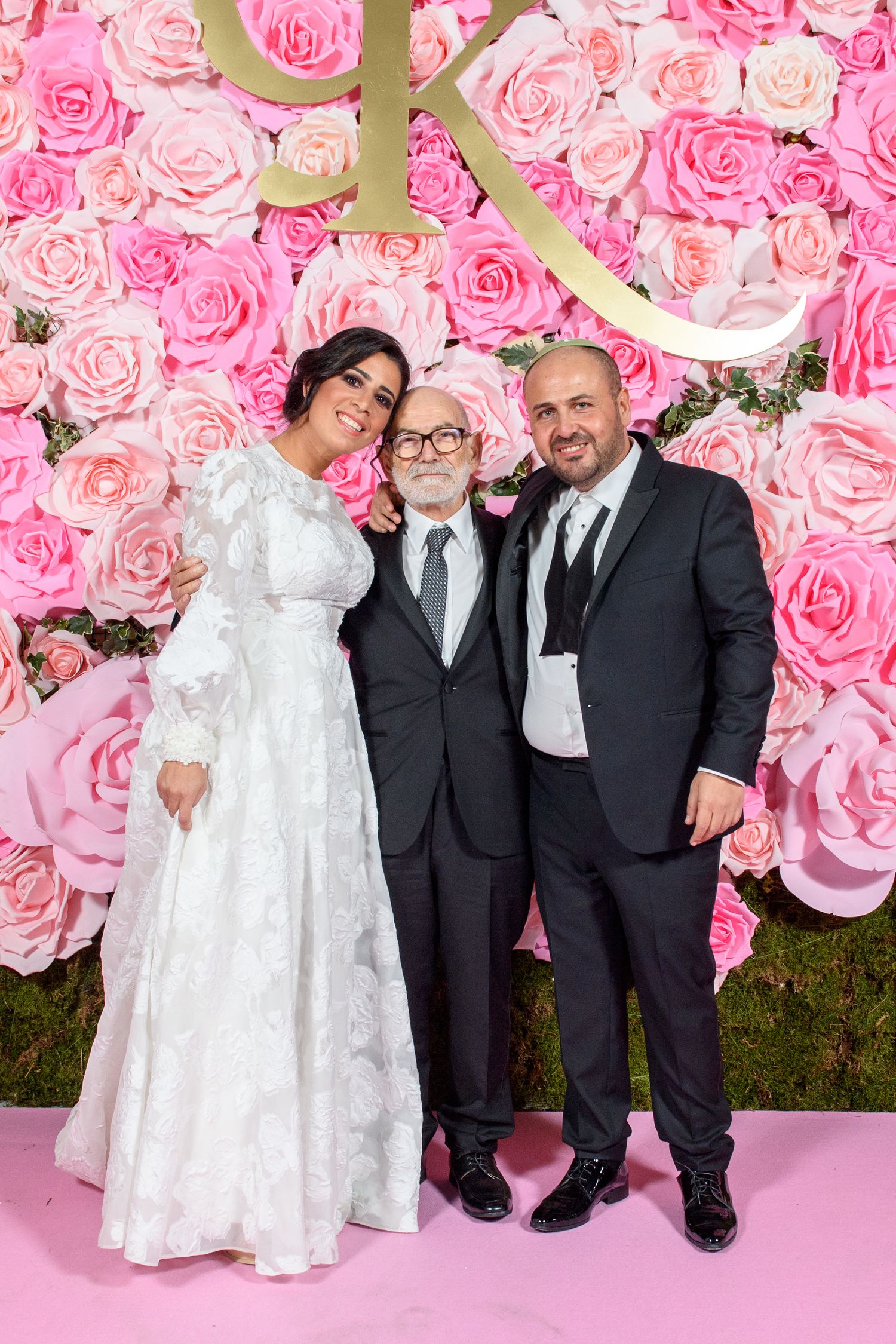 A group poses in formal attire against a pink floral wall backdrop at an elegant event.