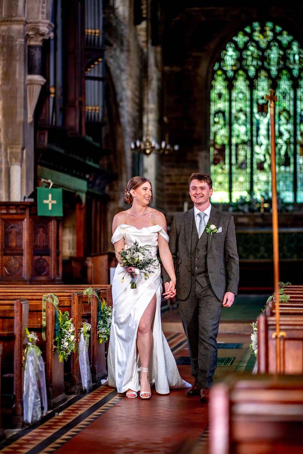 Bride and groom walk hand-in-hand down church aisle, bride in white gown holding bouquet, groom in grey suit.
