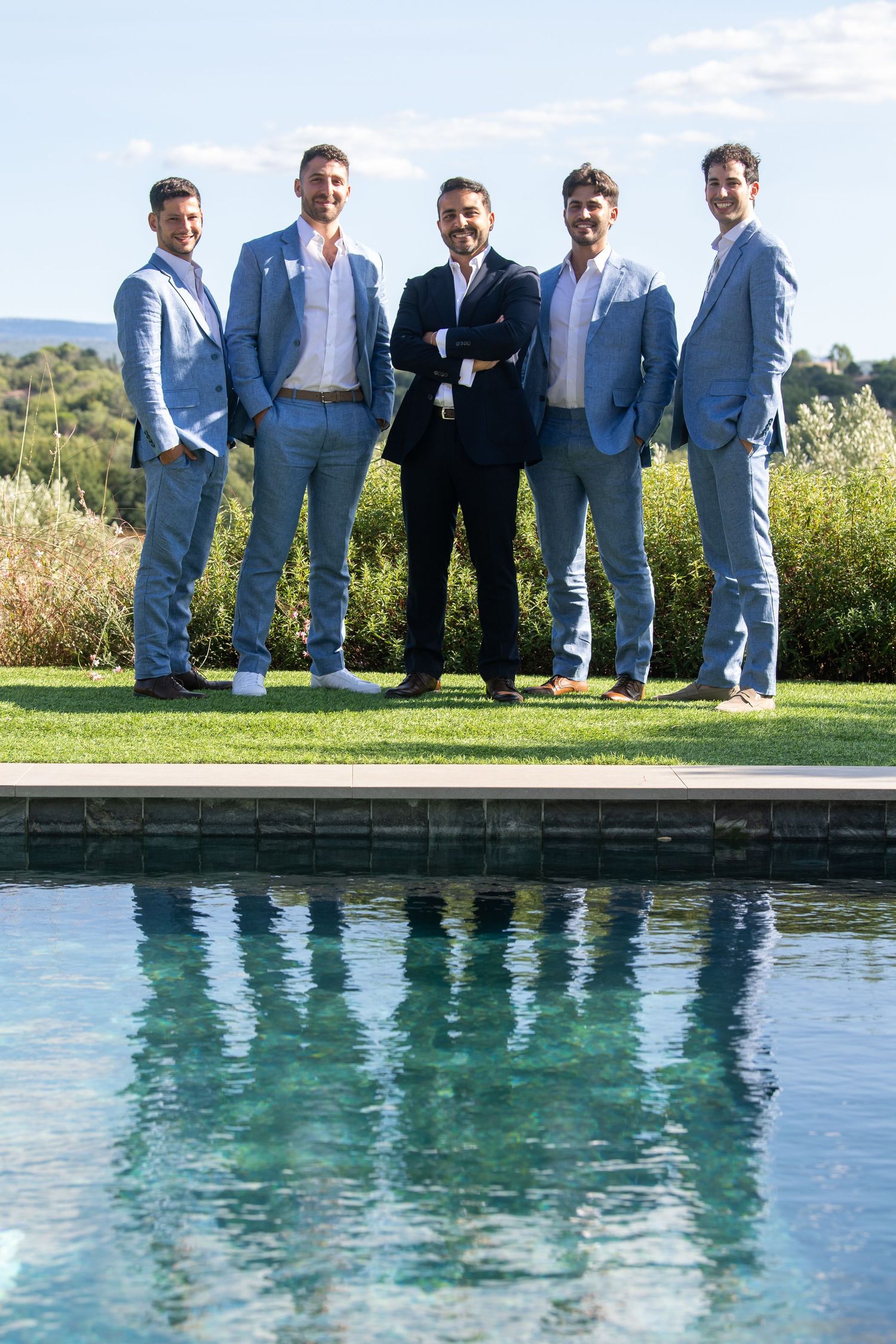 Five men in blue suits pose by a pool outdoors on a sunny day.